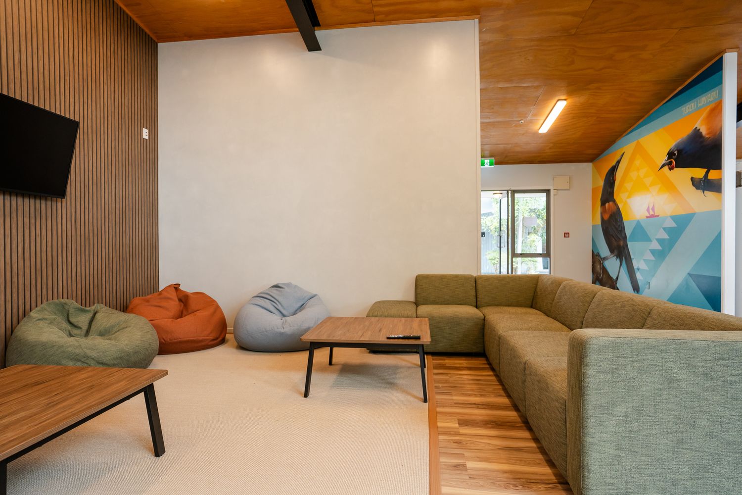 Modern lounge area with colorful bean bags, sectional sofa, wooden ceiling, and vibrant wall mural.