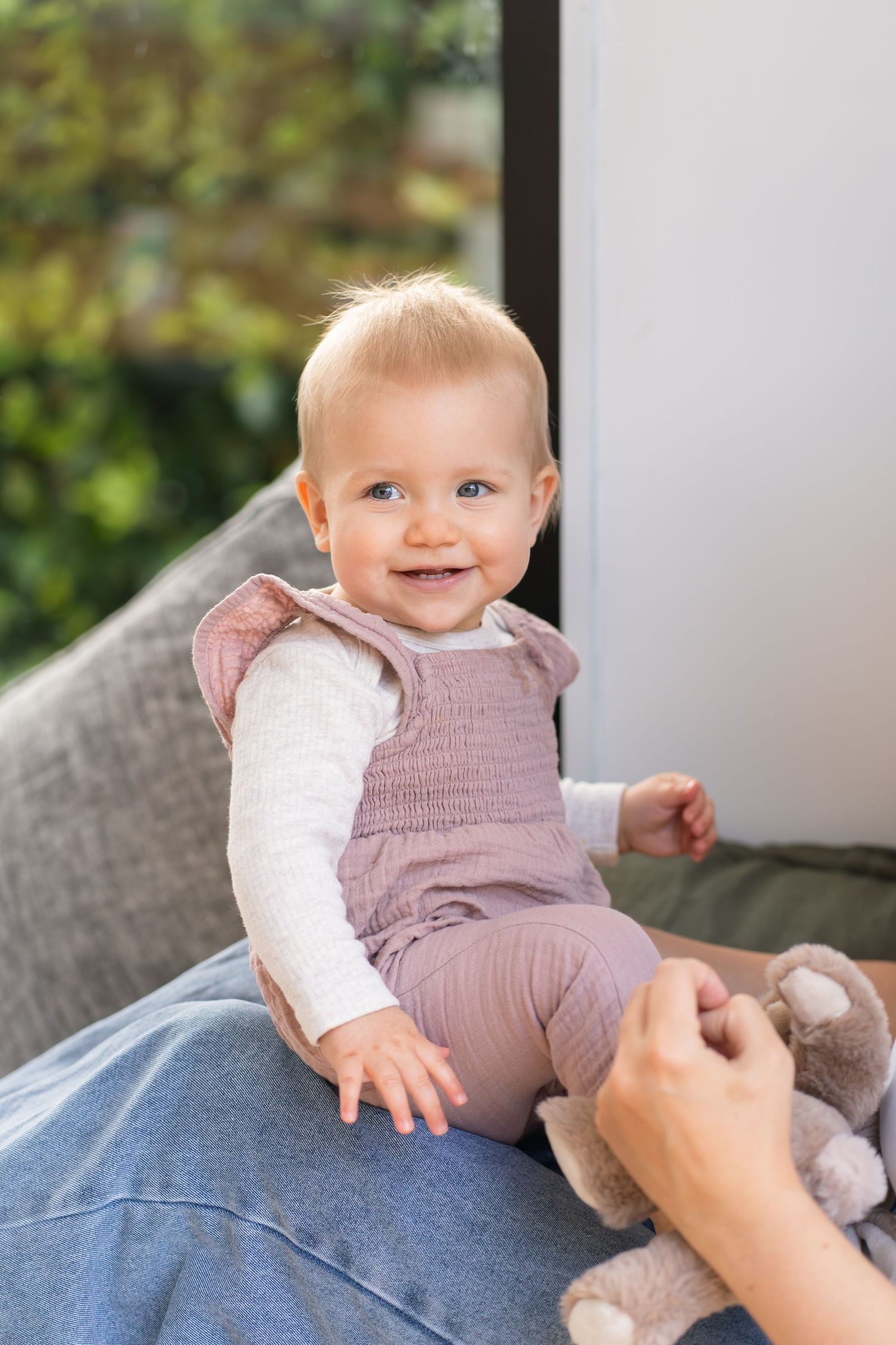 Smiling baby girl in pink outfit sitting on lap with stuffed animal toy near window with green foliage.
