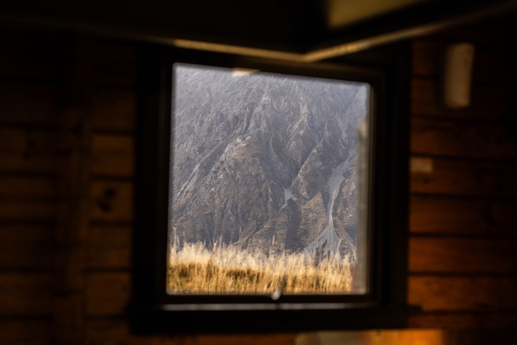 Rustic wooden cabin window framing dramatic mountain cliff and golden grass landscape view outside.