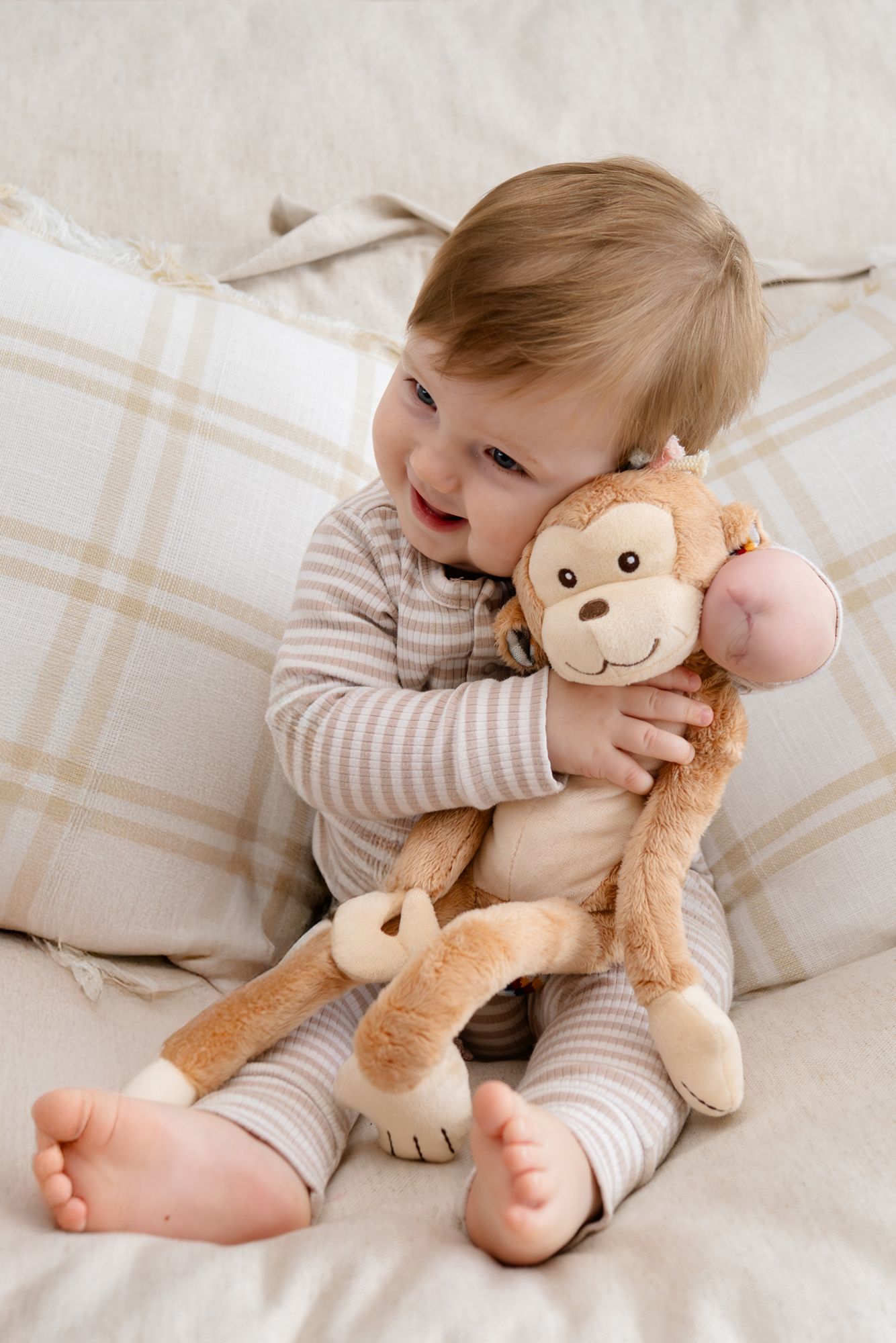 Happy toddler hugging monkey stuffed animal while sitting on beige plaid bedding.