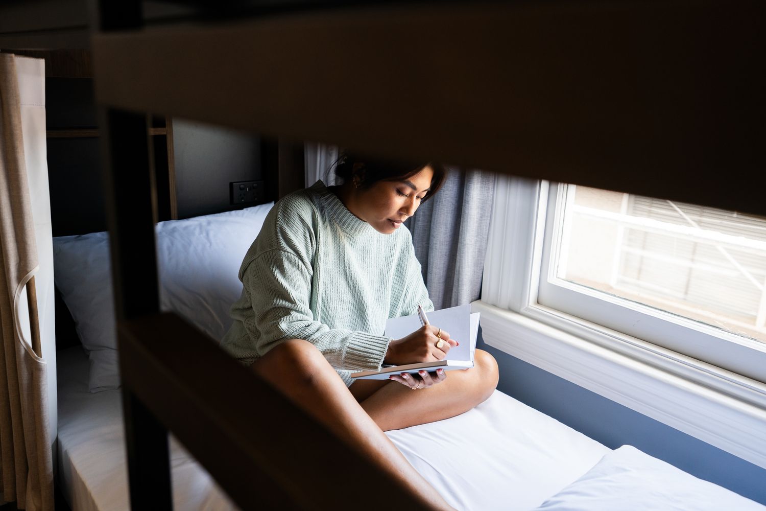 Young woman sitting on a bunk bed writing in a notebook by a window in a hostel room.