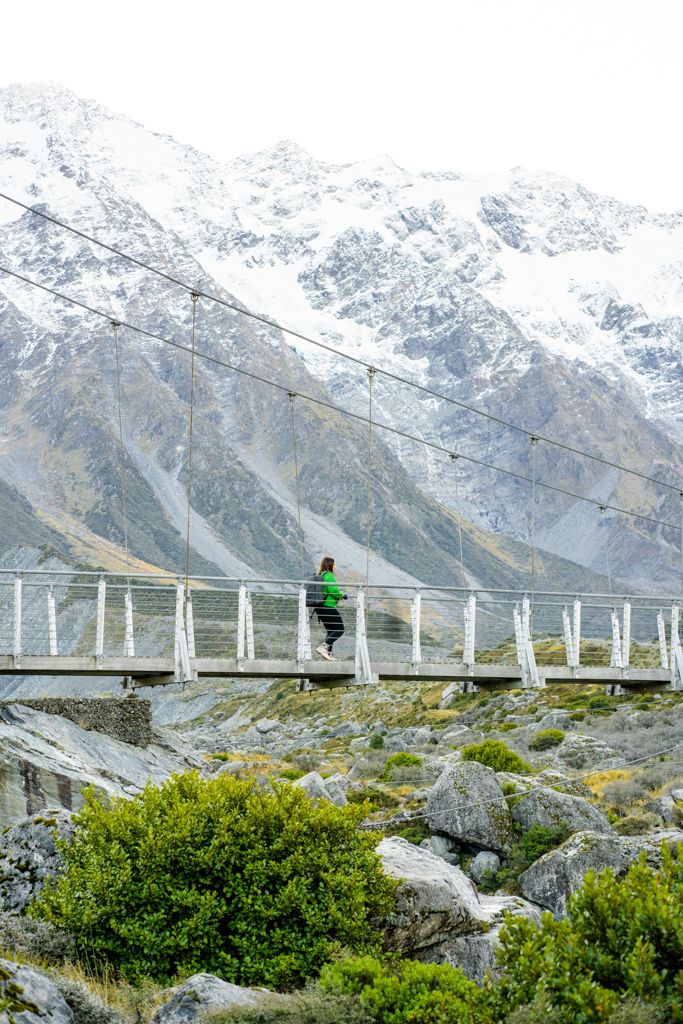 Hiker crossing a suspension bridge with snow-capped mountains in the background on a cloudy day.