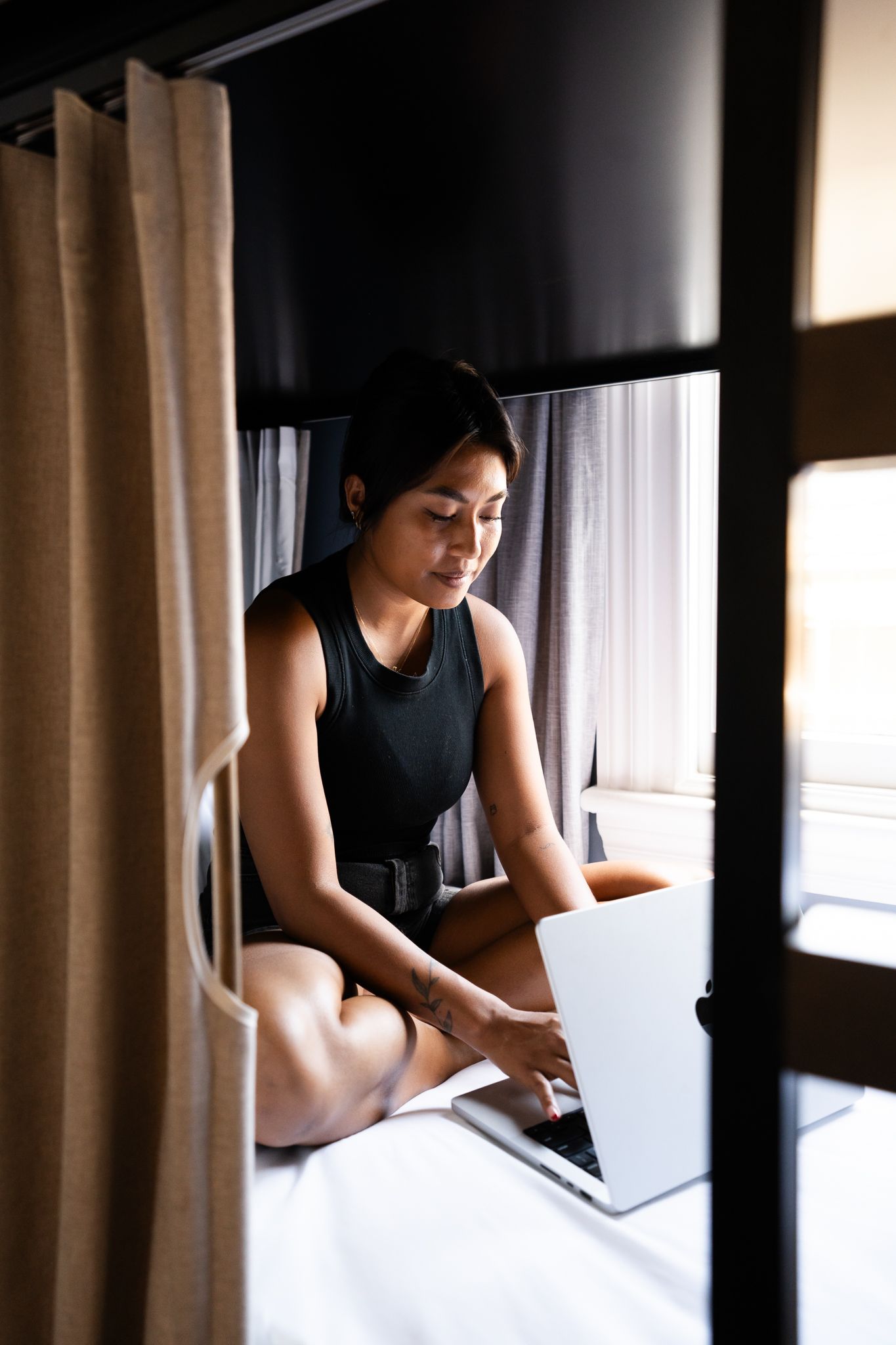 Woman in black outfit working on laptop while sitting cross-legged on a bed in a hostel pod.