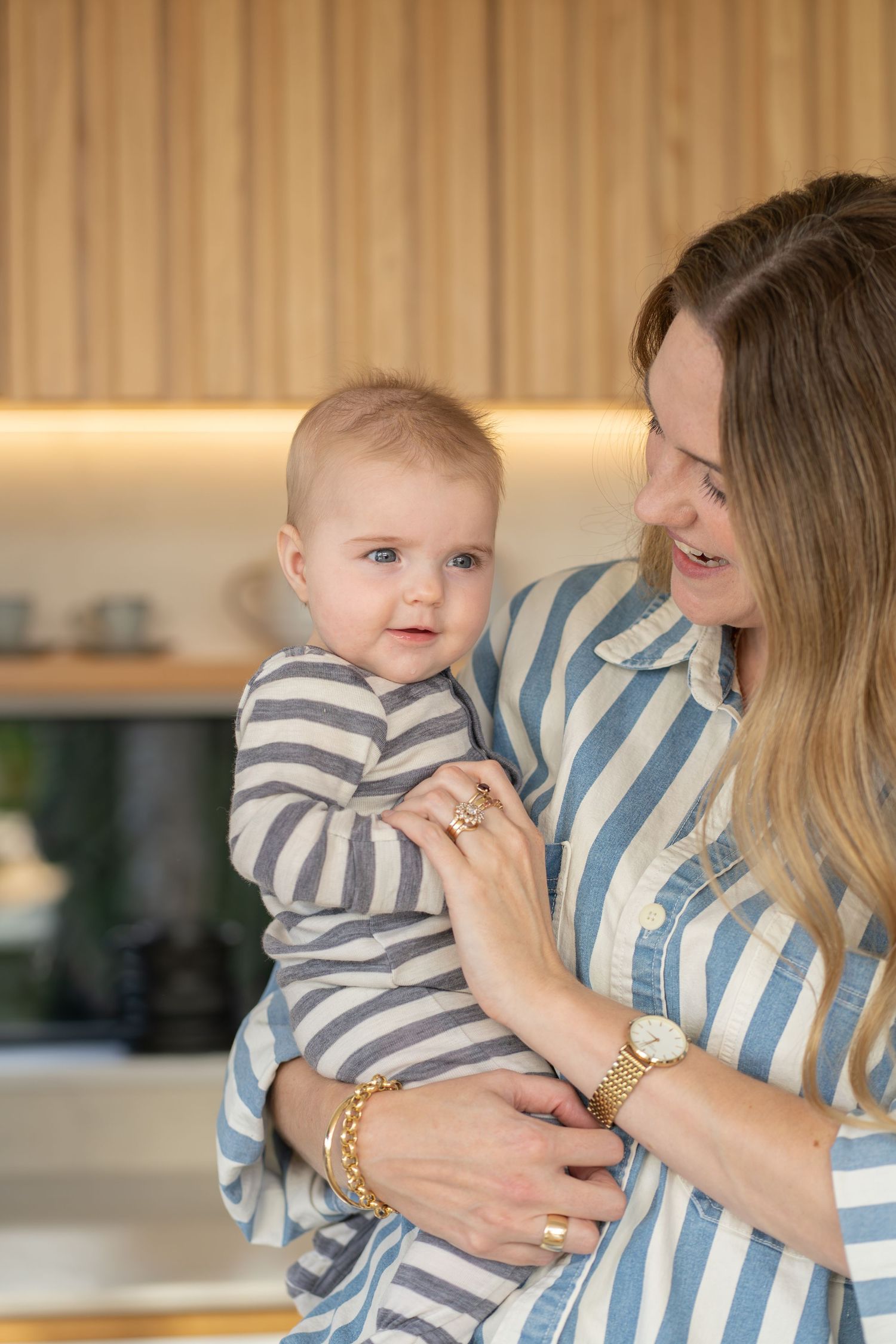 Smiling mother holding her baby in striped outfits in a modern kitchen with wooden accents.