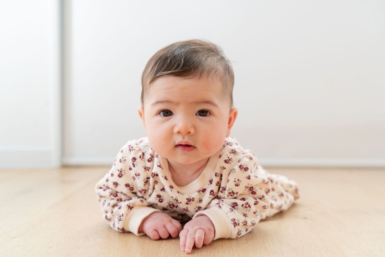 A cute Asian baby in a floral onesie doing tummy time on a wooden floor, looking curiously at the camera.