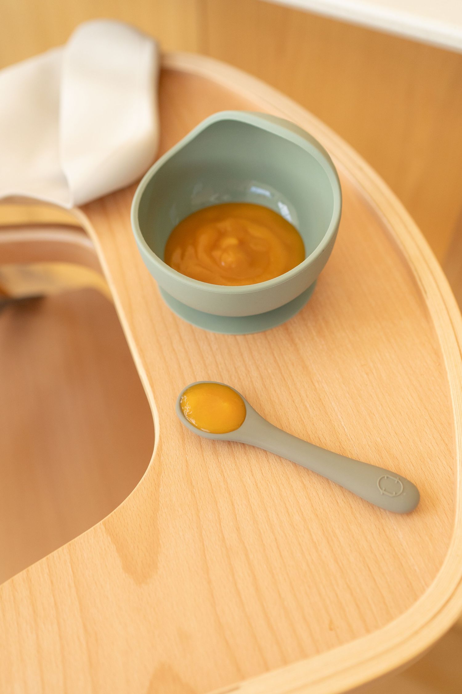 Green silicone baby bowl filled with orange puree and matching spoon on wooden high chair tray.