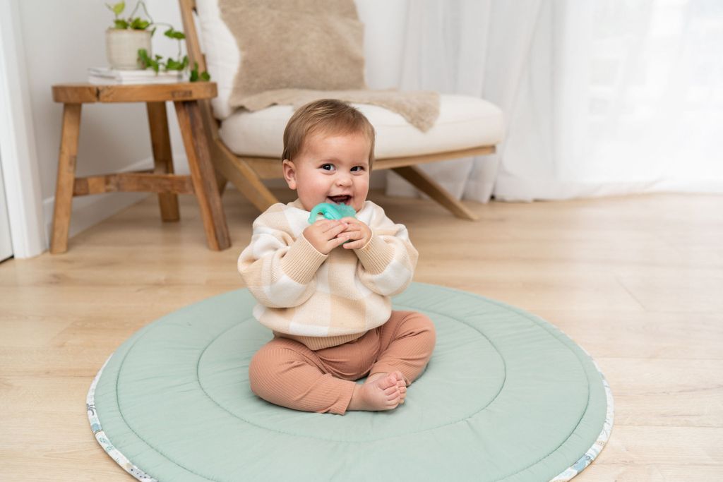 Happy baby sitting on a round green play mat holding a teal teether toy in a bright living room.