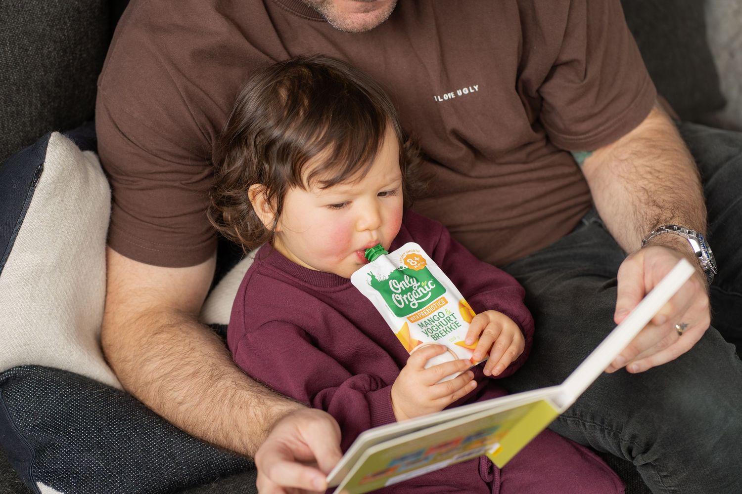 A father reads a colorful book to his toddler, who sips from a green baby food pouch while sitting on a cozy couch.
