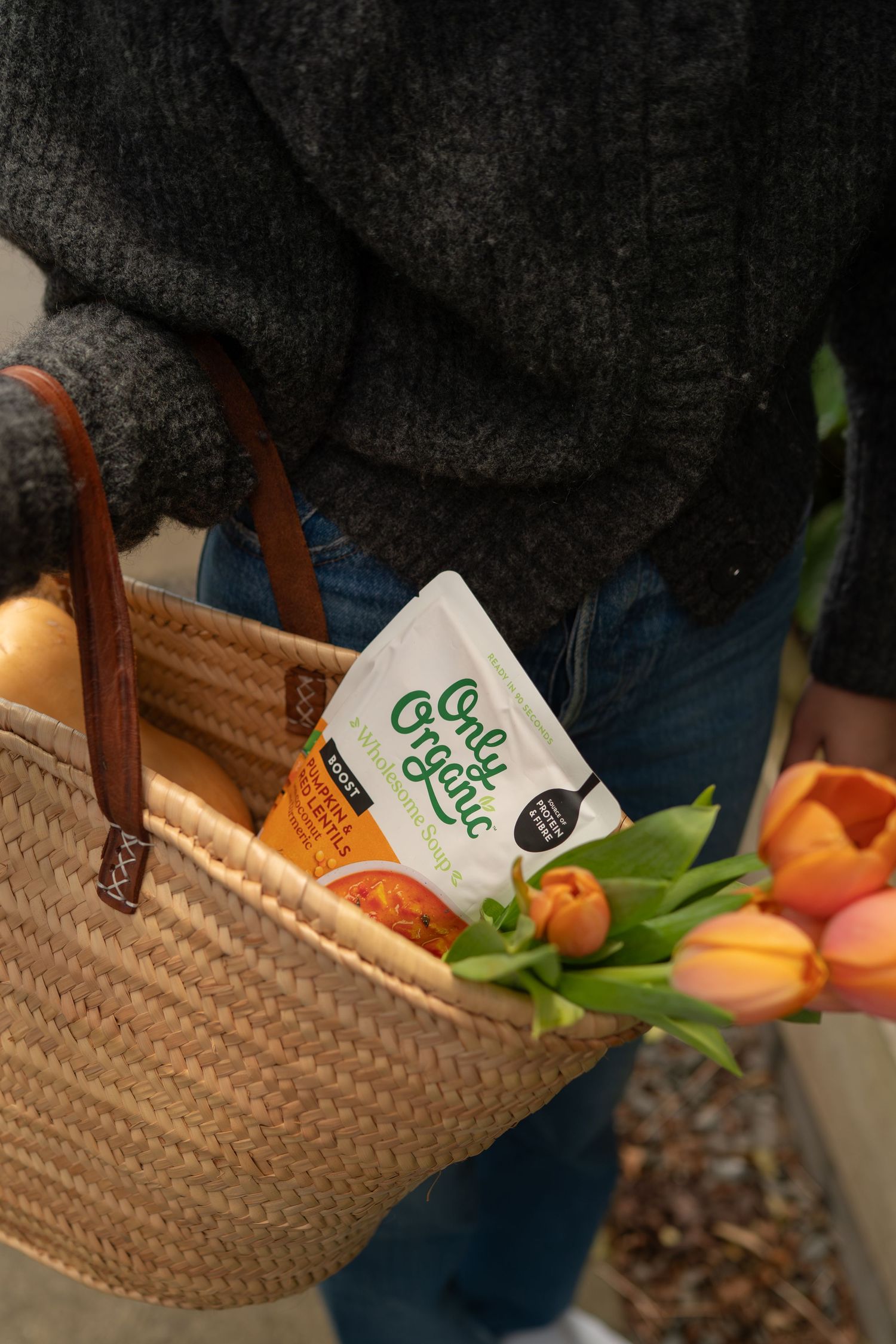 Person carrying a wicker tote bag with Onle Organic product, bread, and fresh orange tulips.