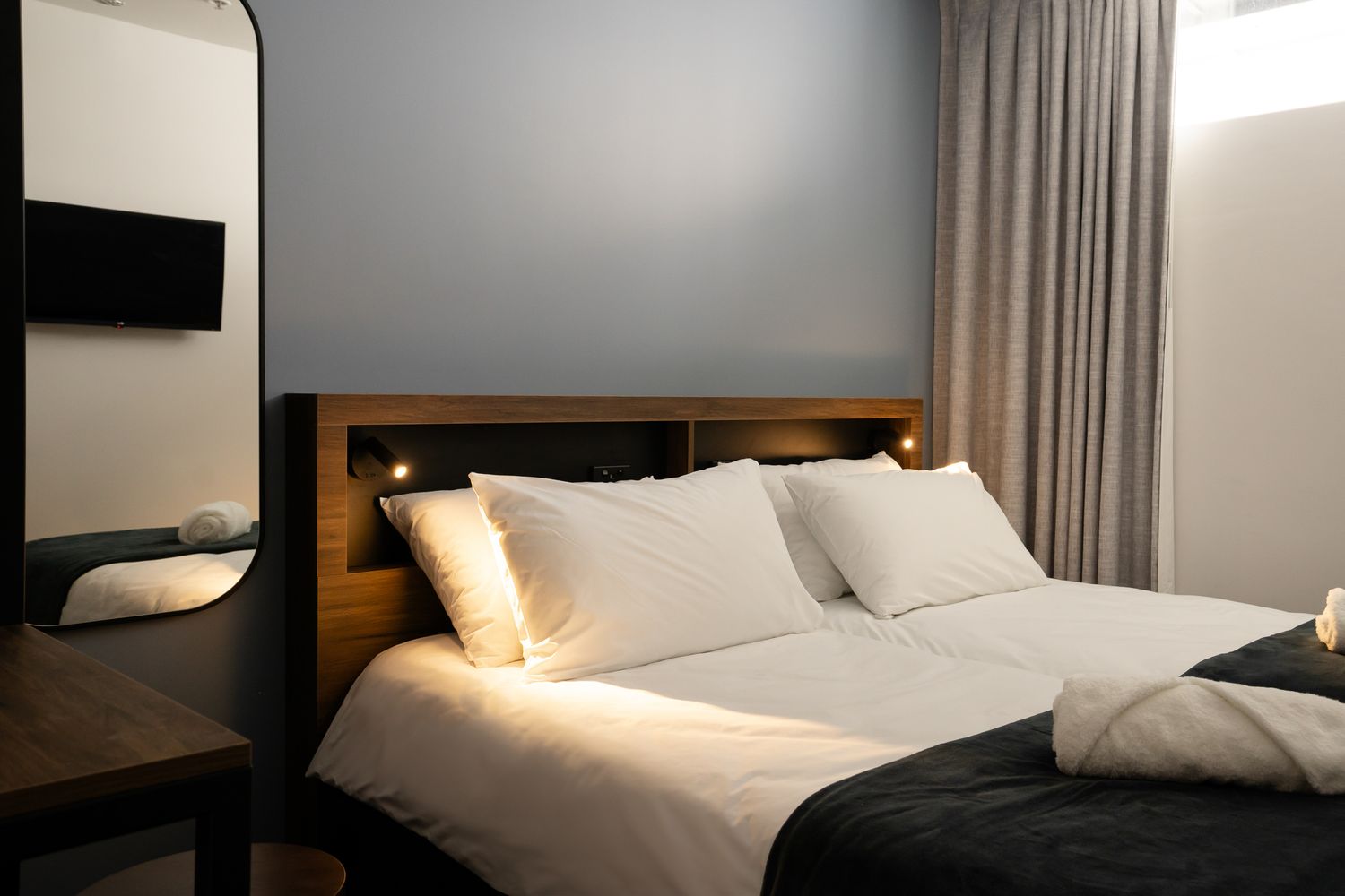 Cozy hotel room with a neatly made double bed, white pillows, wooden headboard and soft lighting.