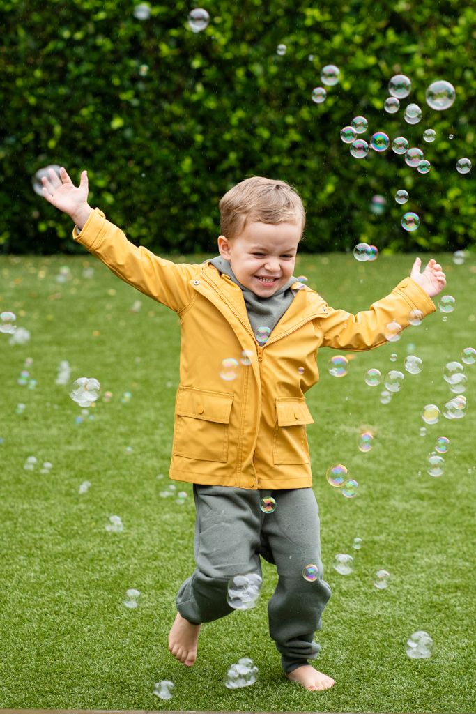 Happy young boy in yellow raincoat playing with soap bubbles in a green garden.