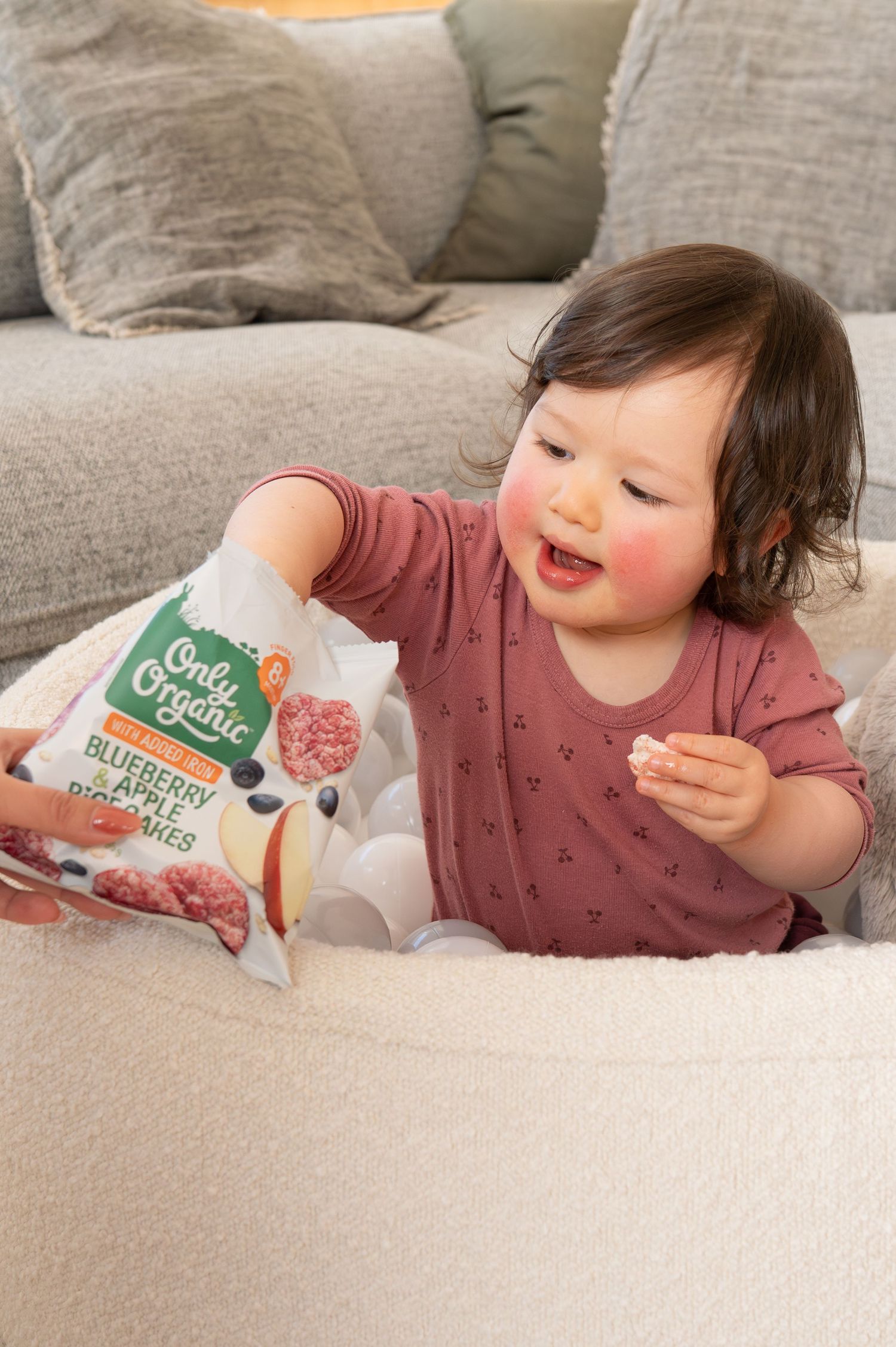 Smiling toddler in pink shirt holding Only Organic Blueberry Apple Rice Cakes bag while sitting in a ball pit near a couch.