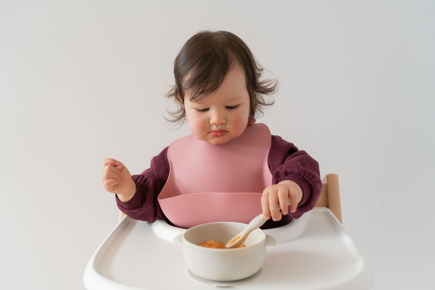Cute baby girl in pink bib sitting in high chair eating food with spoon from white bowl.