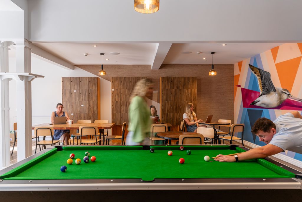 Colorful hostel common room with green pool table, pendant lights, wooden walls, and guests socializing in background.
