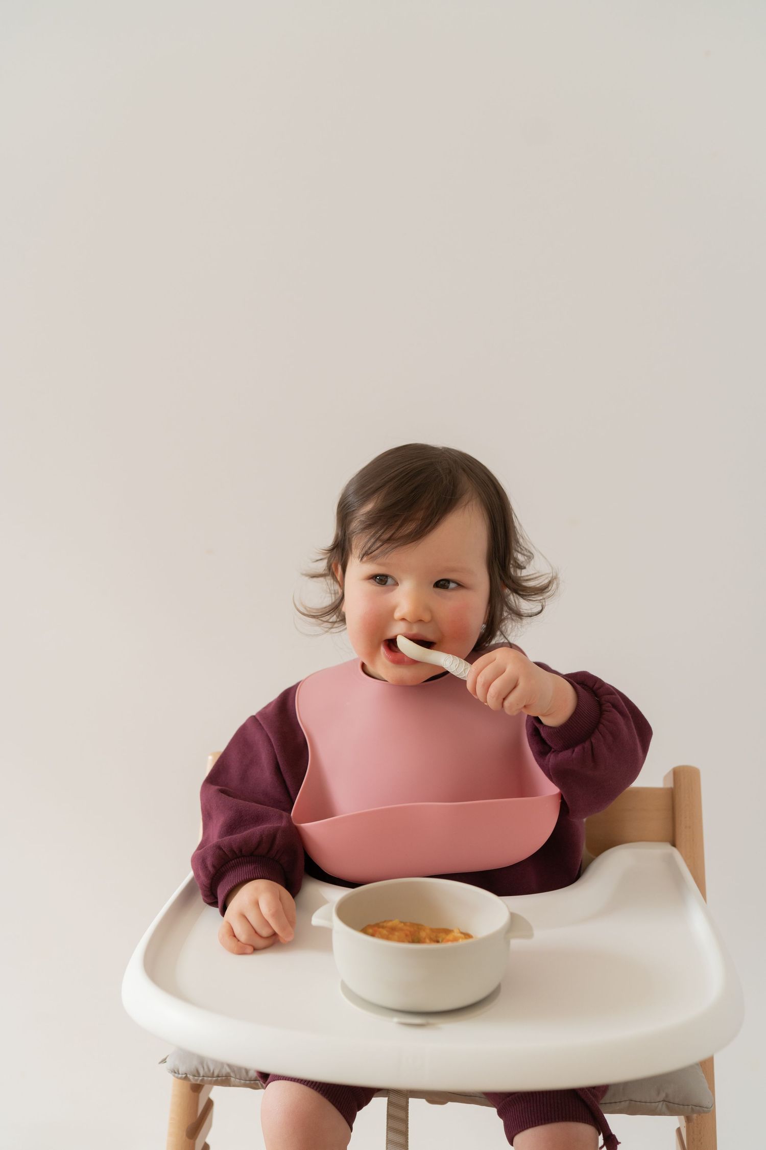 Baby in high chair wearing pink bib, eating with spoon from bowl, dressed in burgundy top.