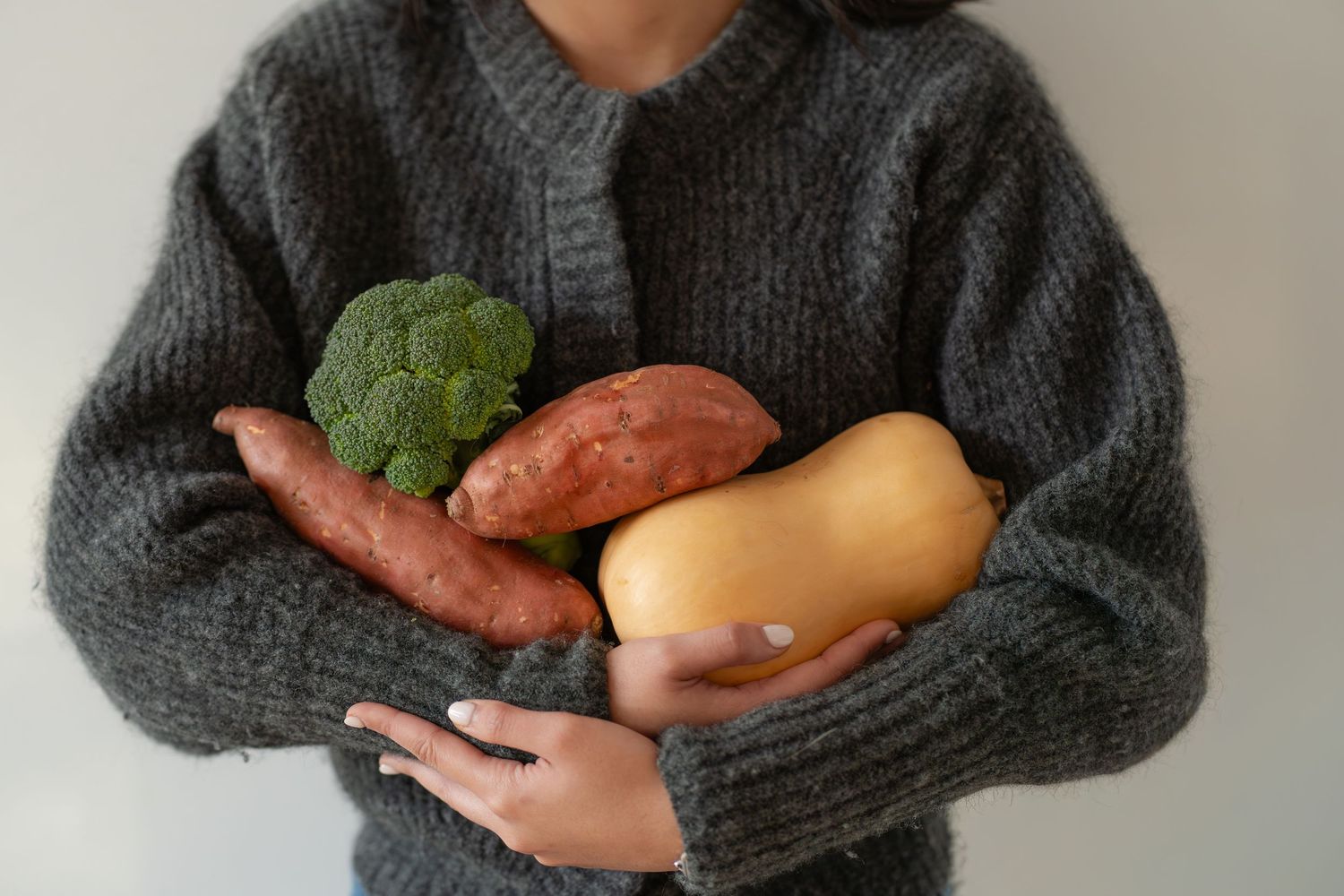 Person in gray knit sweater holding fresh vegetables including broccoli, sweet potatoes, and butternut squash.