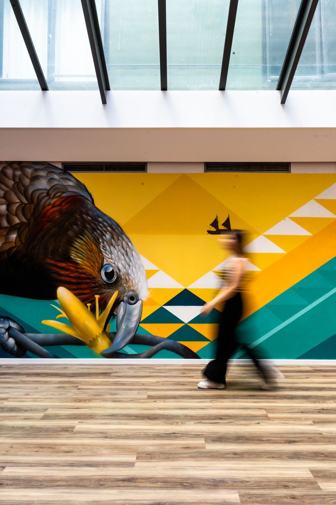Person walking past a large colorful bird mural in a modern building with skylights.