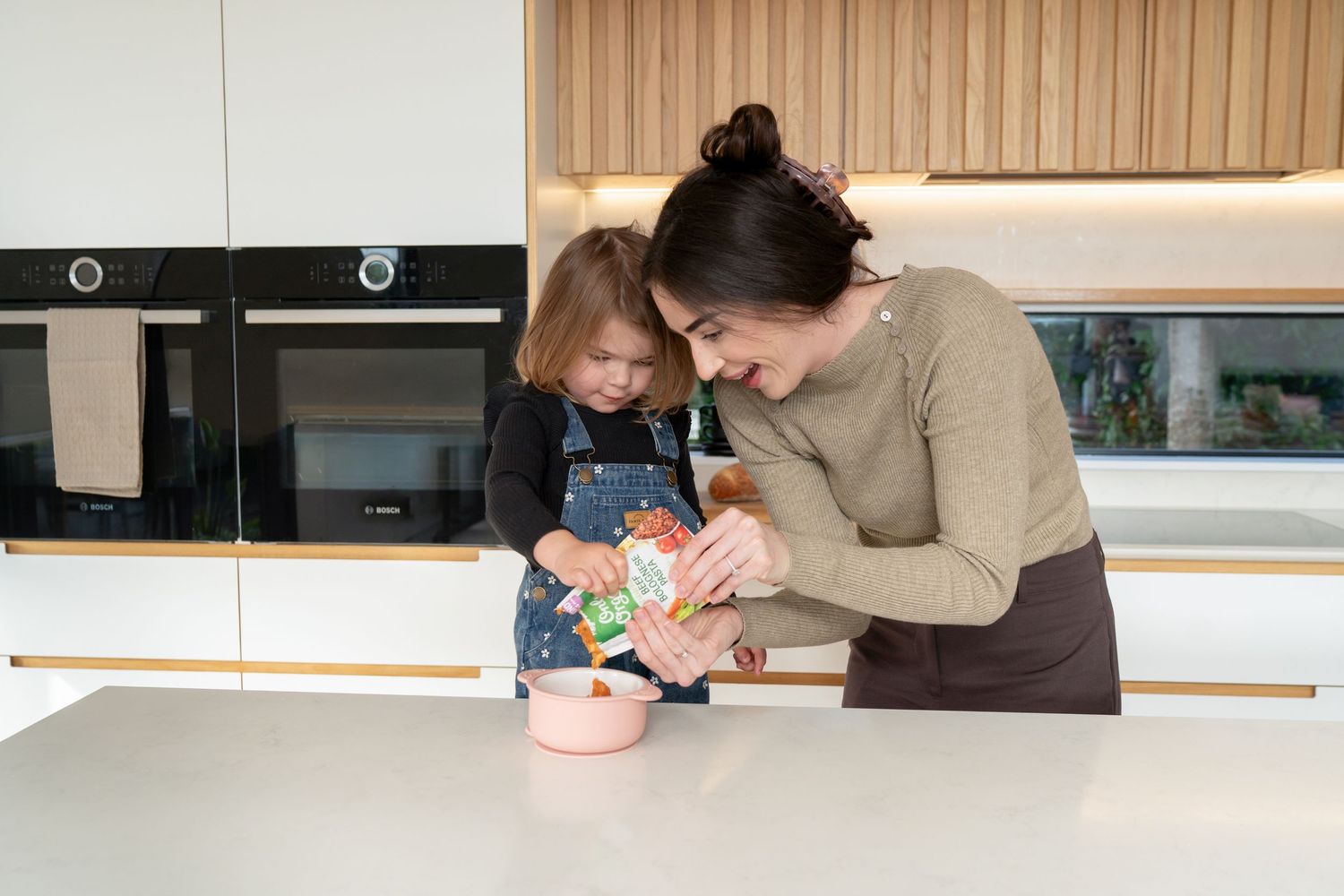 A mother and young daughter cook together in a modern white kitchen, adding ingredients to a pot.