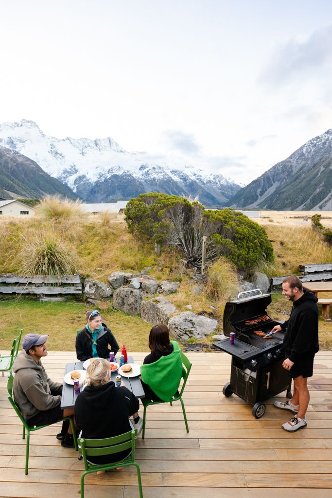 Group of friends enjoying a BBQ on a wooden deck with stunning snow-capped mountain views in New Zealand.