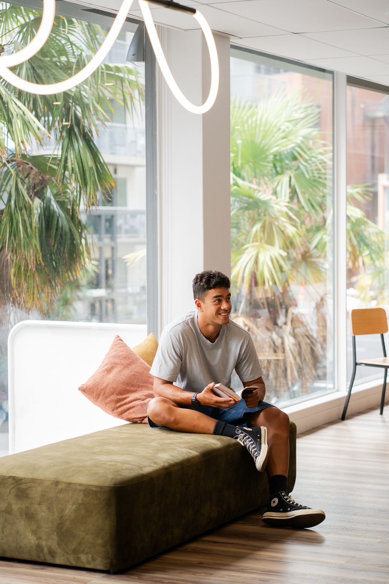 Smiling young man sitting on a green couch holding a phone in a modern hostel lounge with palm trees.