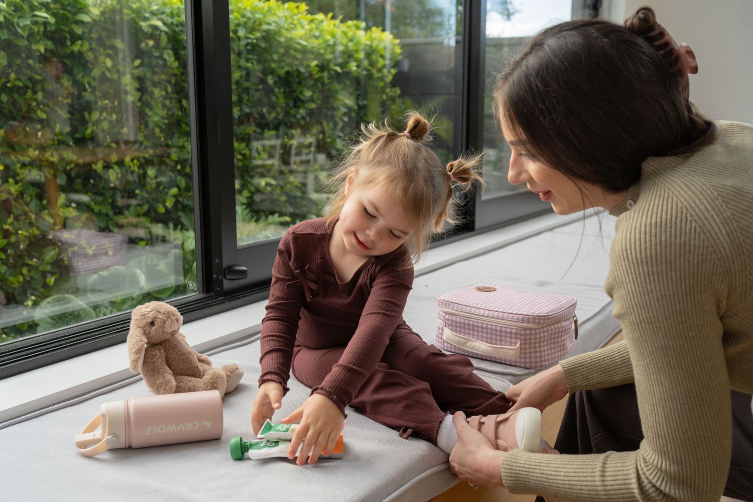 A mother and young daughter sit by a large window, playing together with toys and a pink storage box indoors.