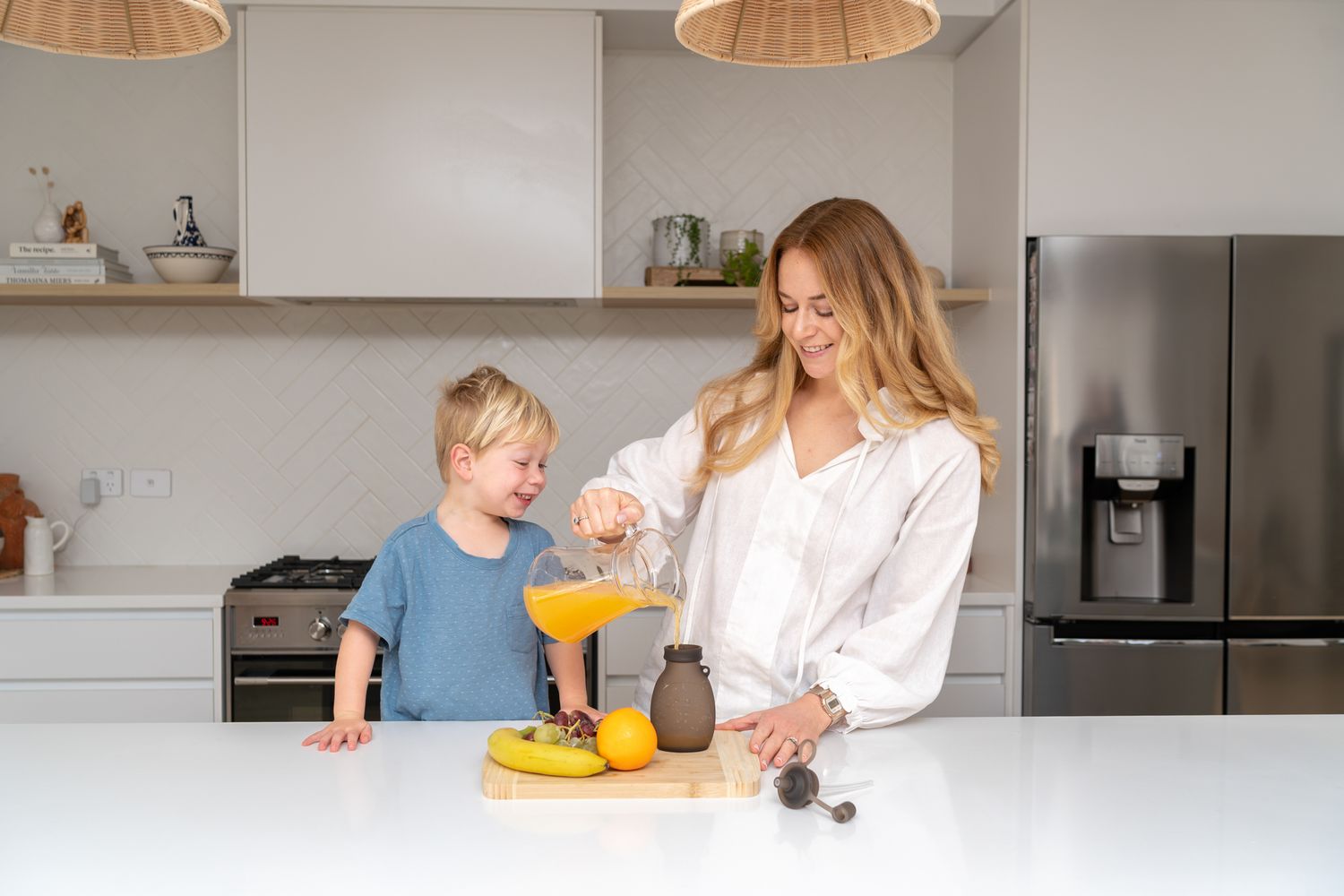 Smiling mother and young child preparing fresh fruit juice together in modern white kitchen.