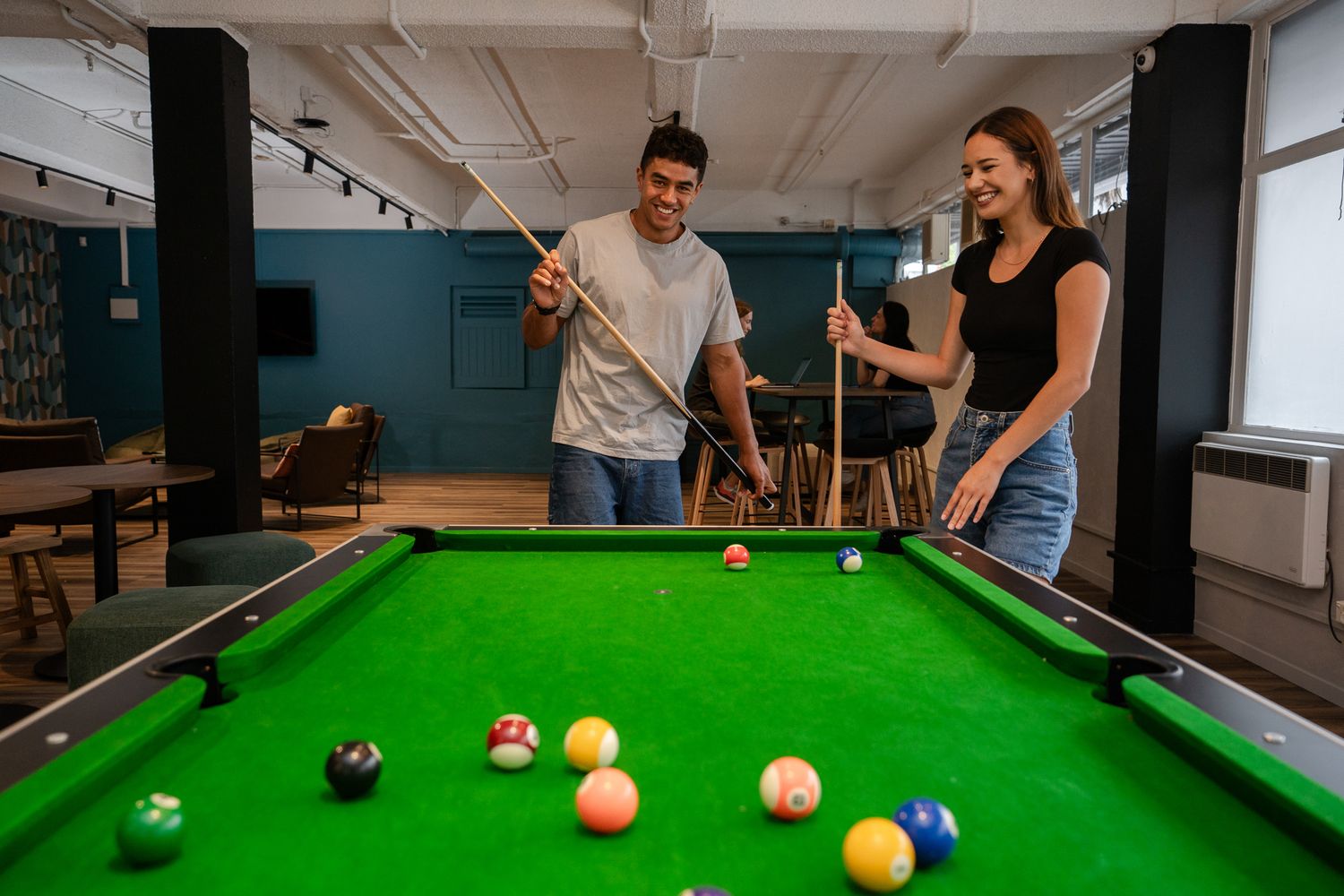 Young couple playing pool together in a vibrant hostel common room with green billiard table.