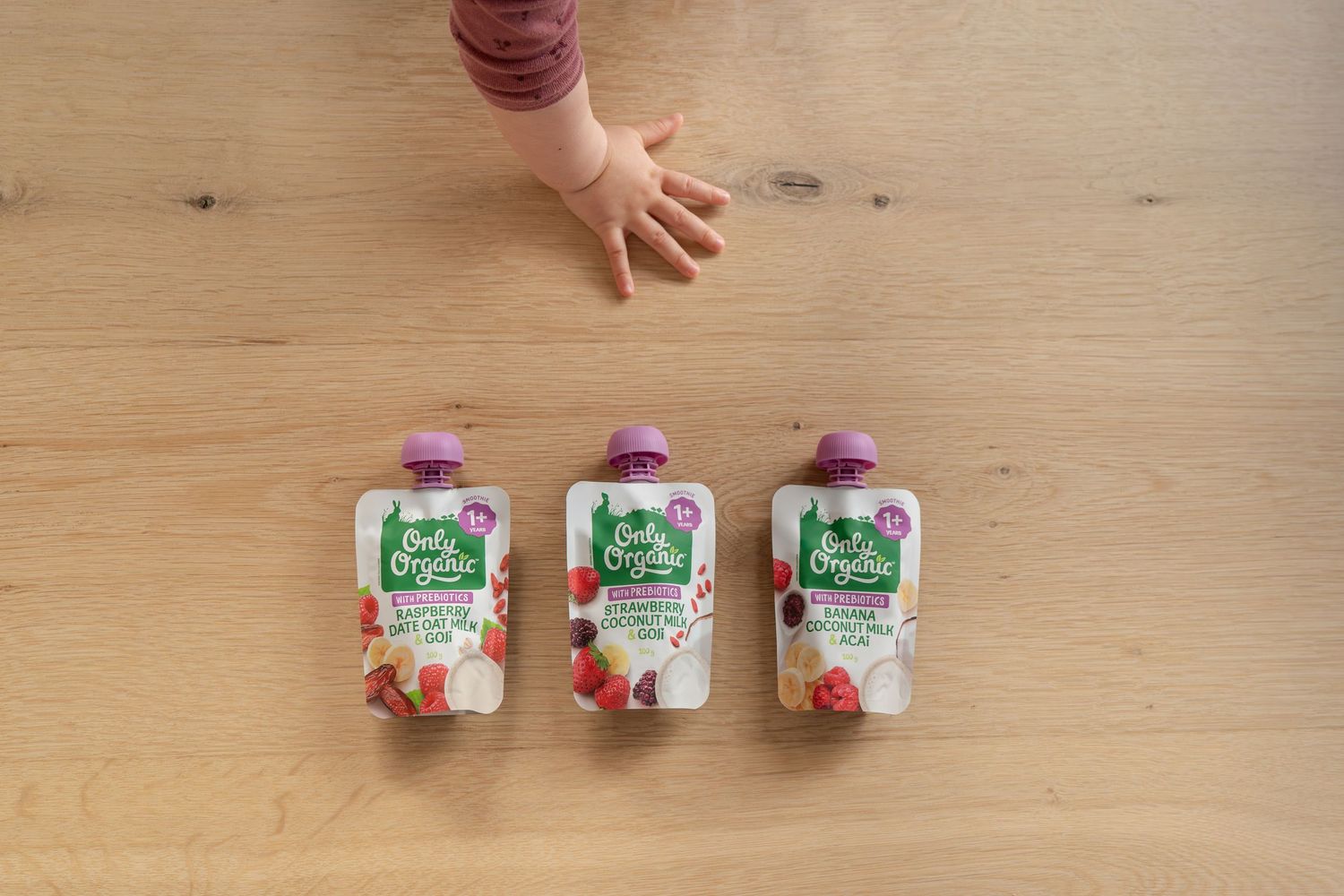 Three Ella's Organic baby food pouches on a wooden table with a baby's hand reaching toward them.