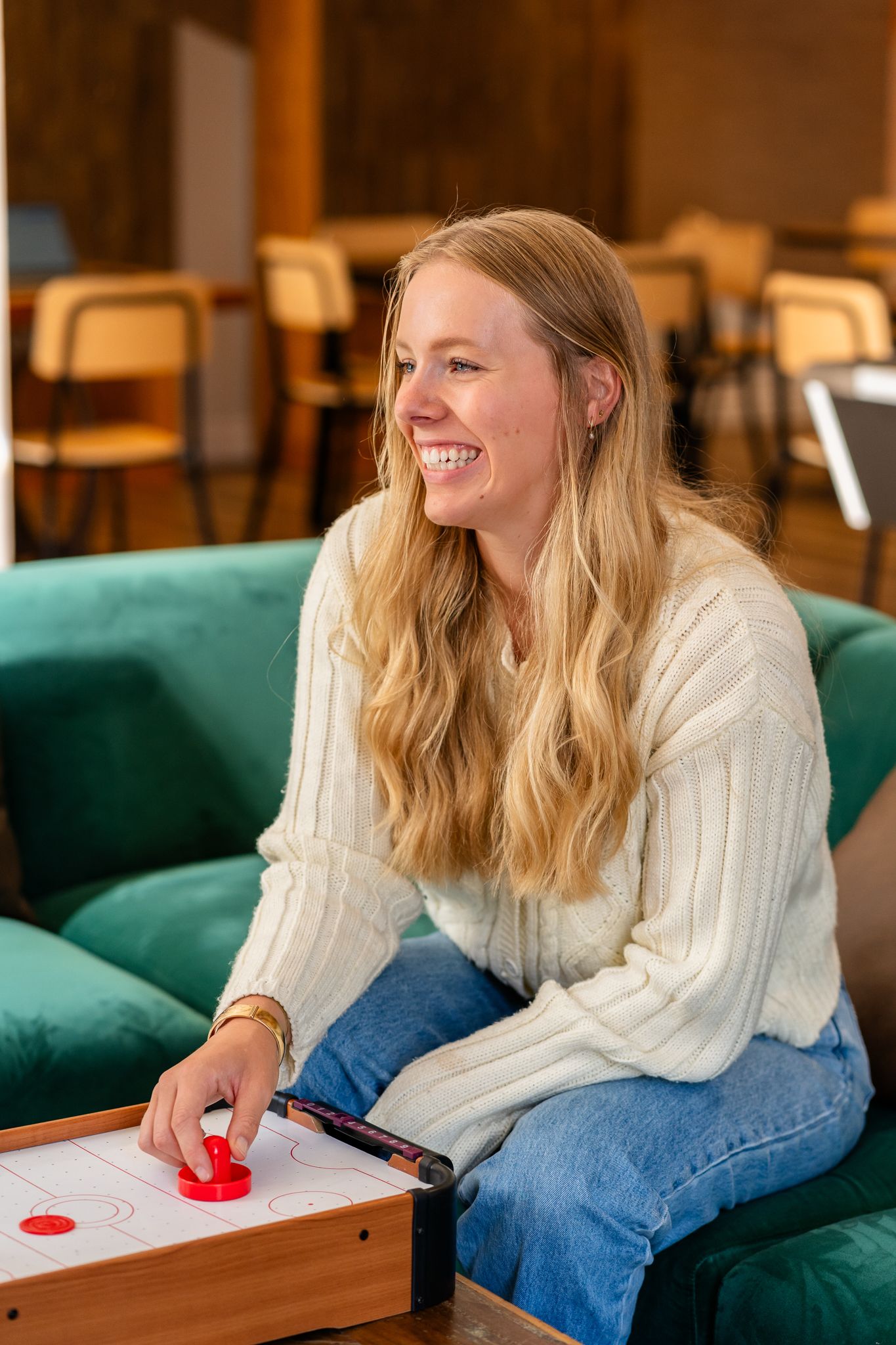 Smiling blonde woman playing tabletop air hockey on a green sofa in a hostel lounge.