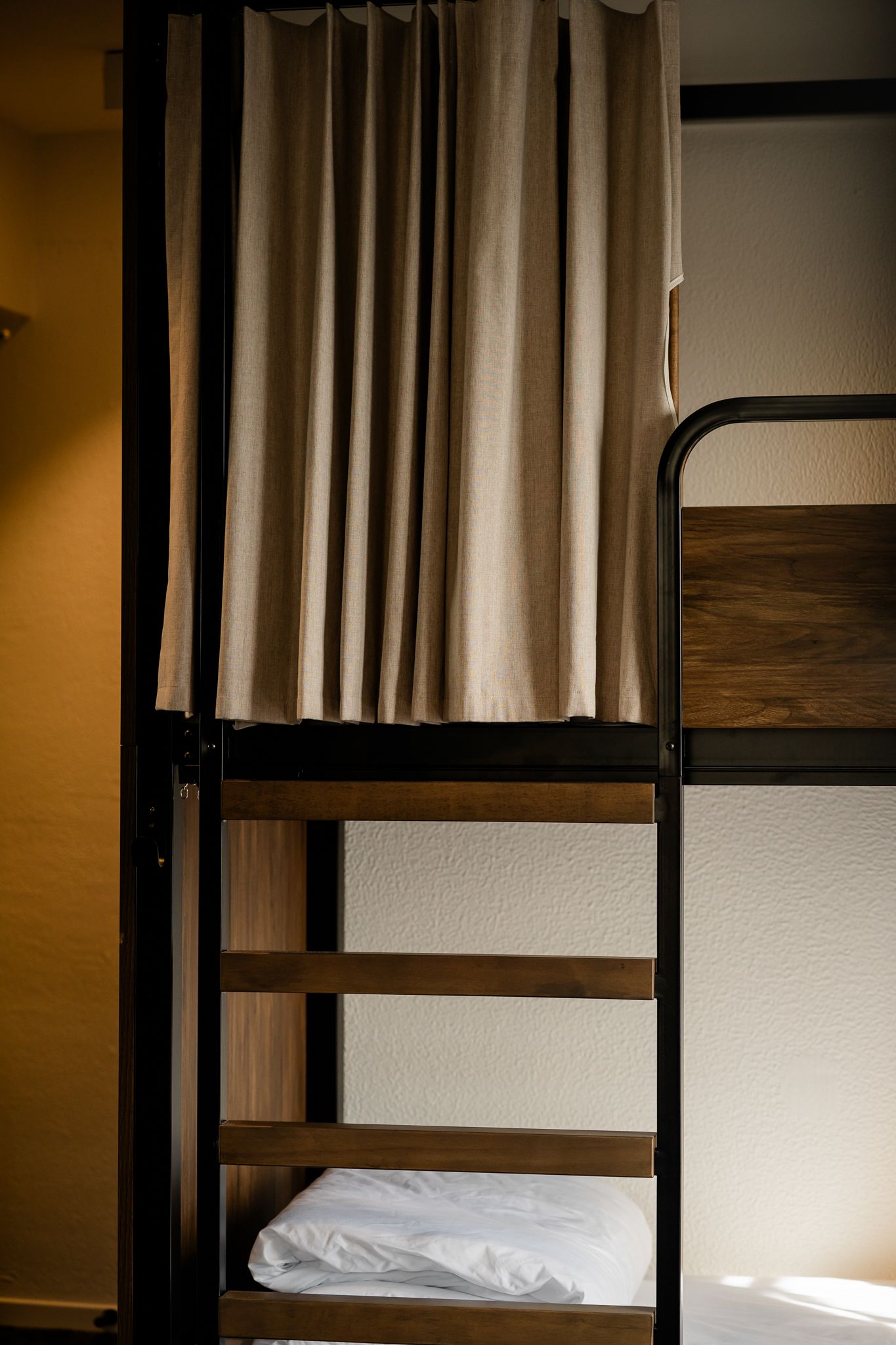 Modern bunk bed with wooden frame, black metal ladder, and privacy curtain in a hostel dormitory.