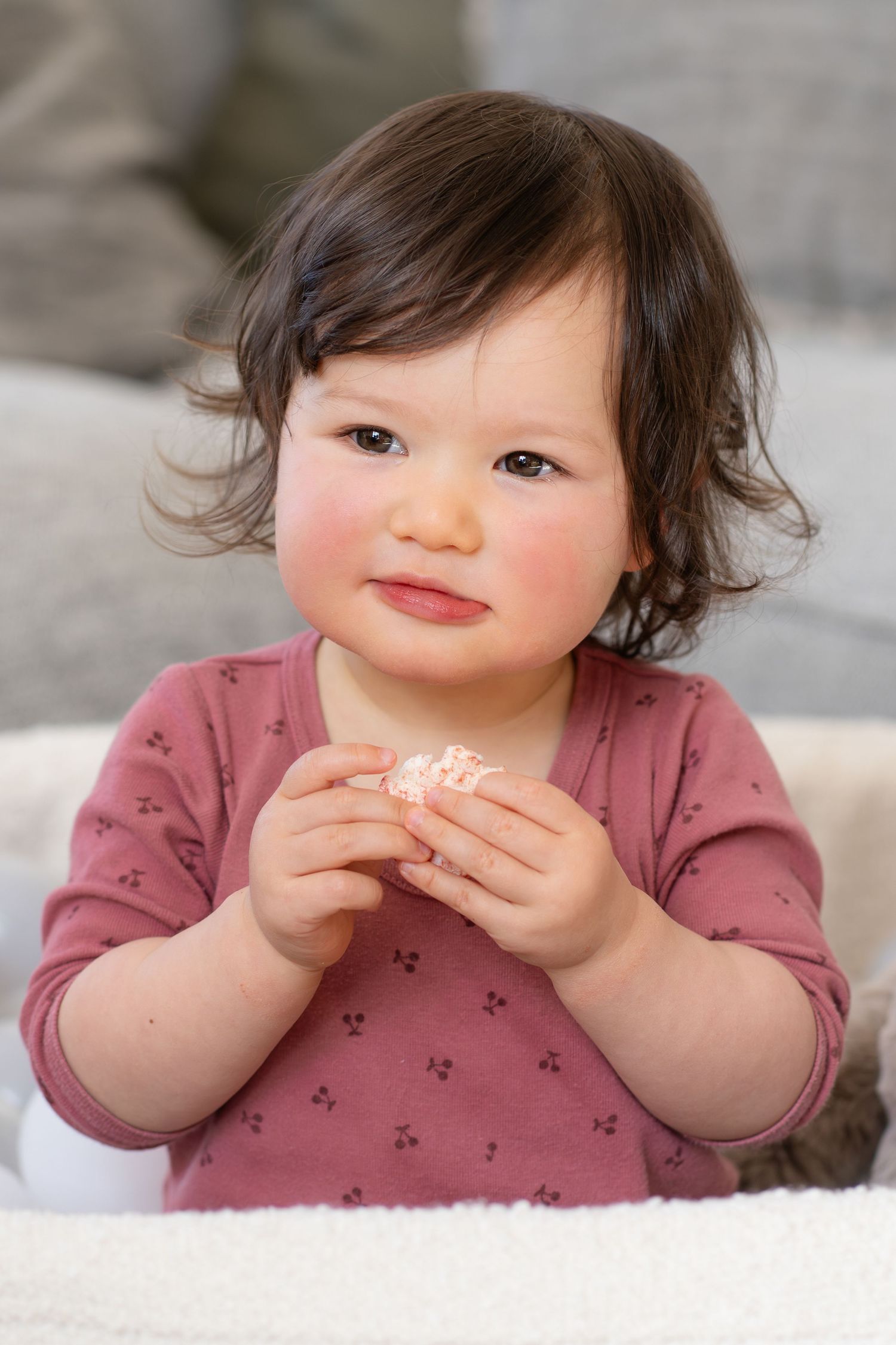 Adorable toddler girl with dark curly hair wearing a pink floral top, holding a small snack while sitting on a white fluffy blanket.