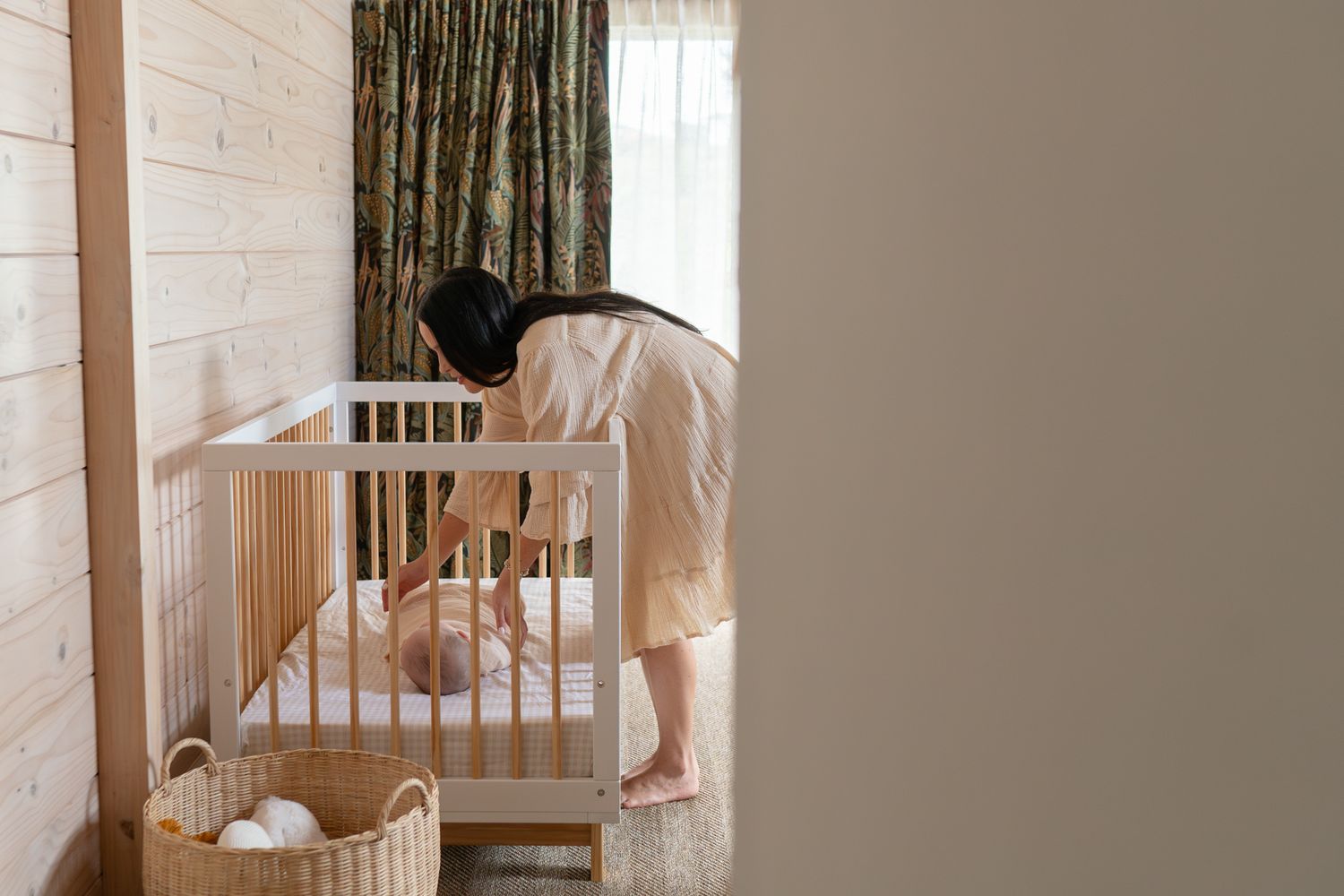 Mother leaning over white wooden crib tending to baby in a cozy nursery room.