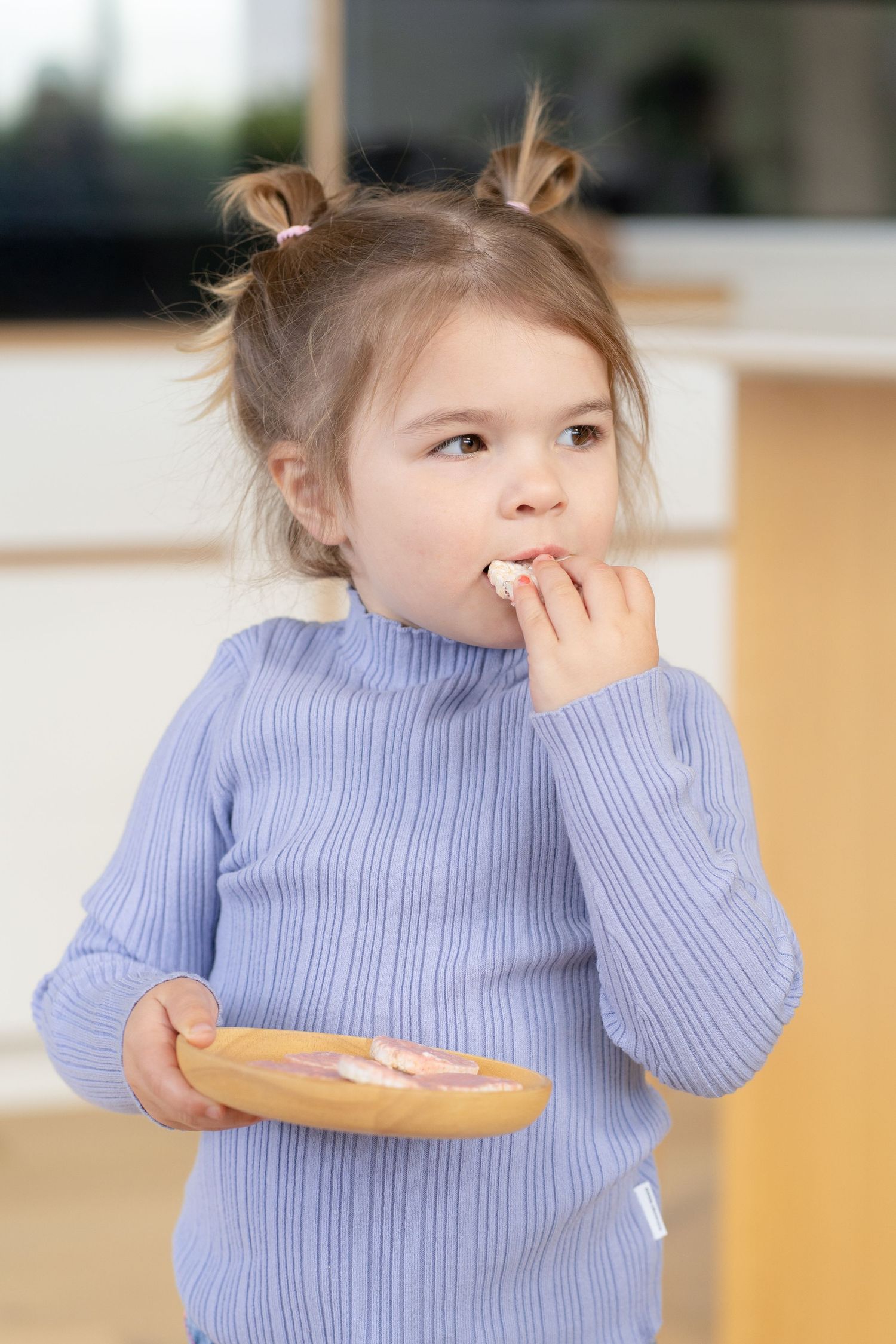 Young girl in blue ribbed sweater eating snack from wooden plate, standing in bright kitchen.