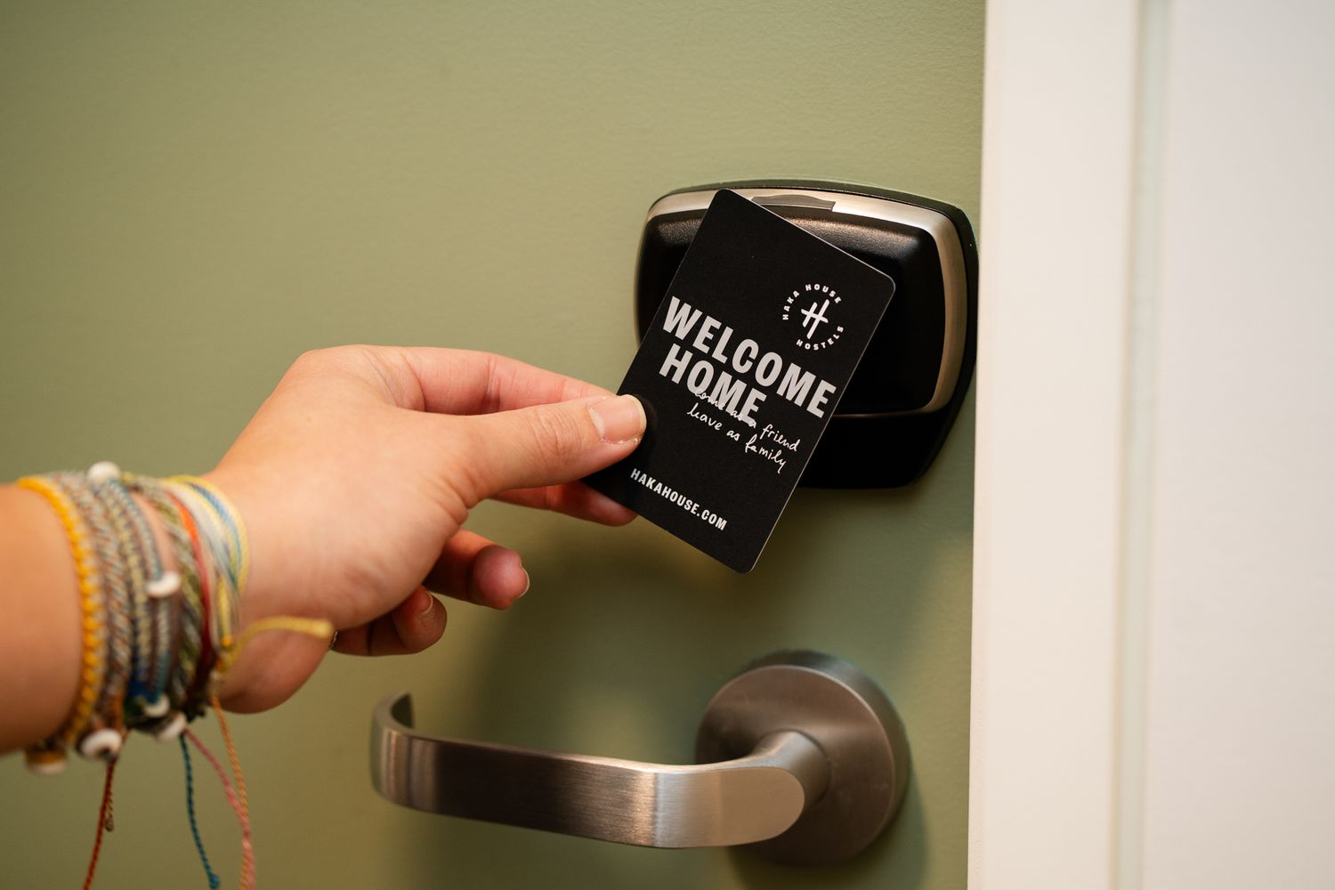 Hand holding a Welcome Home keycard against a modern hotel door lock with green wall background.