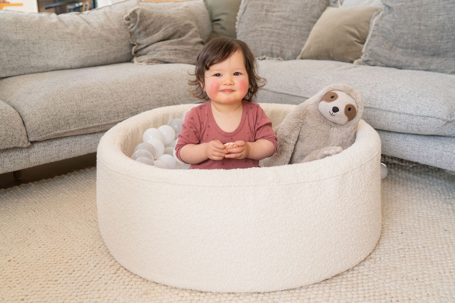 Smiling baby sitting in a round white foam play pit with a plush sloth toy on a living room carpet.