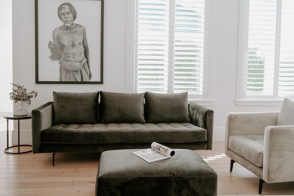 Bright minimalist living room with olive velvet sofa, ottoman, and large portrait artwork above.