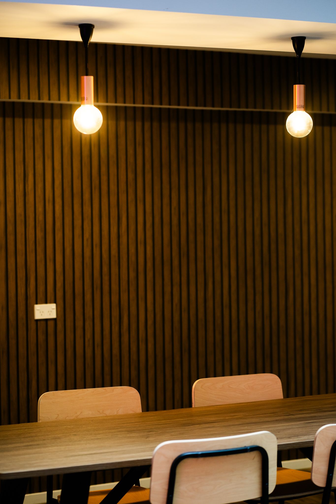 Modern dining room with pendant bulb lights hanging over a long wooden table with chairs against slatted wood wall.