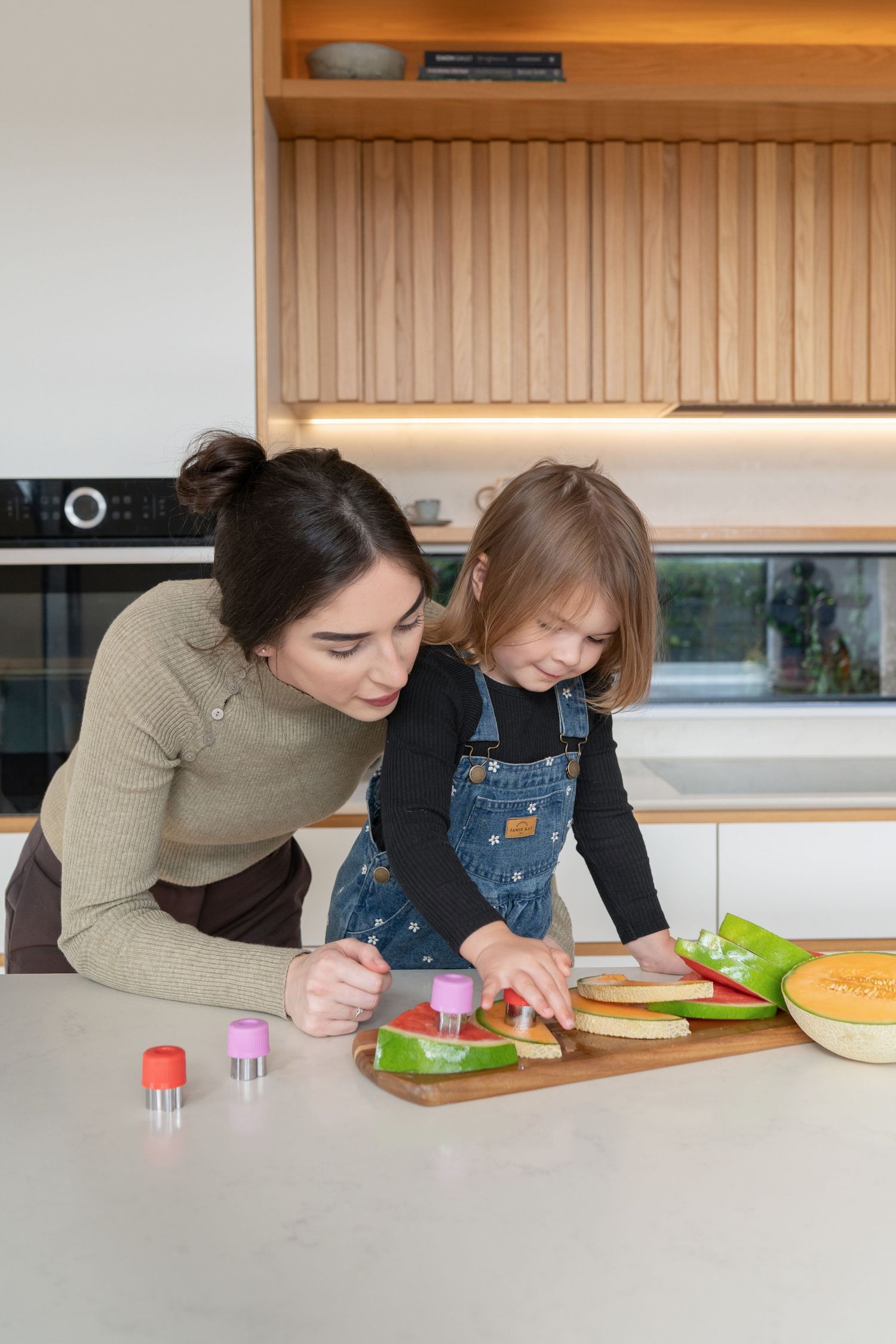 A young mother and her little girl use cookie cutters to prepare fruit snacks together at a modern kitchen counter.