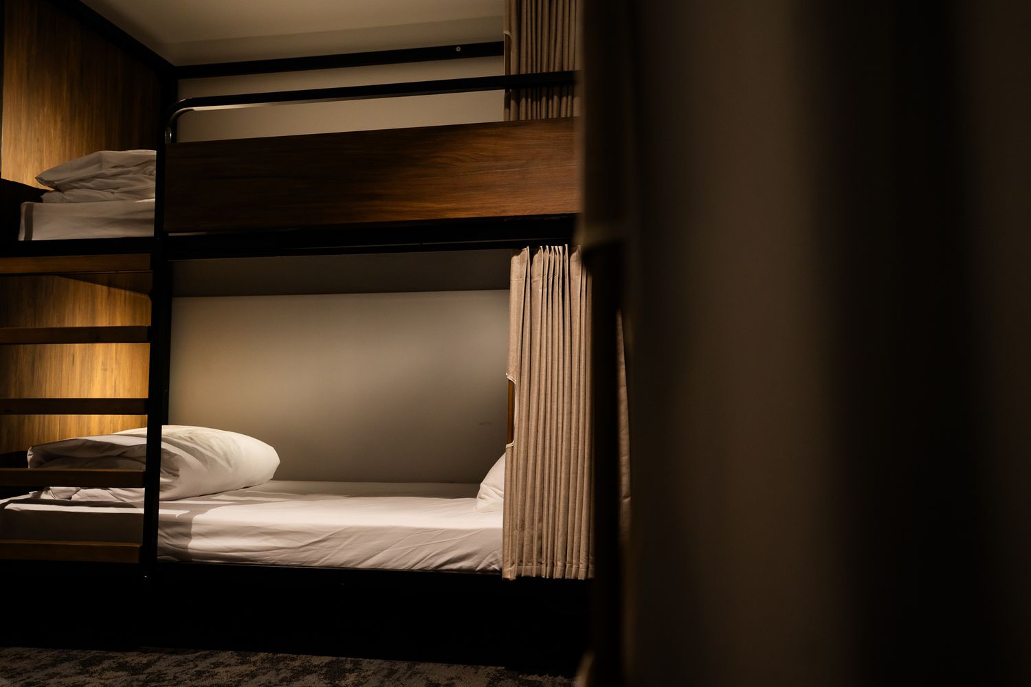 Minimalist hostel bunk beds with privacy curtains and wooden paneling in a dark cozy dormitory room.
