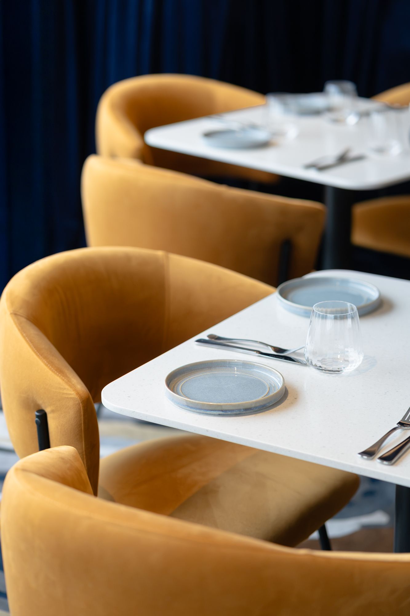 Modern restaurant interior with mustard yellow velvet chairs and white tables set with plates.