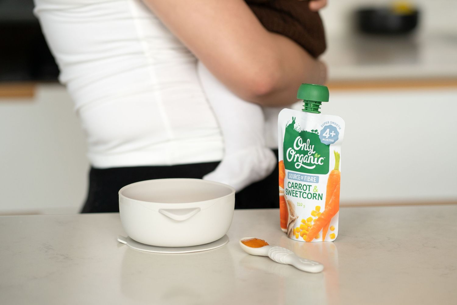 Only Organic carrot sweetcorn baby food pouch beside a bowl and spoon on a kitchen counter with a parent in background.