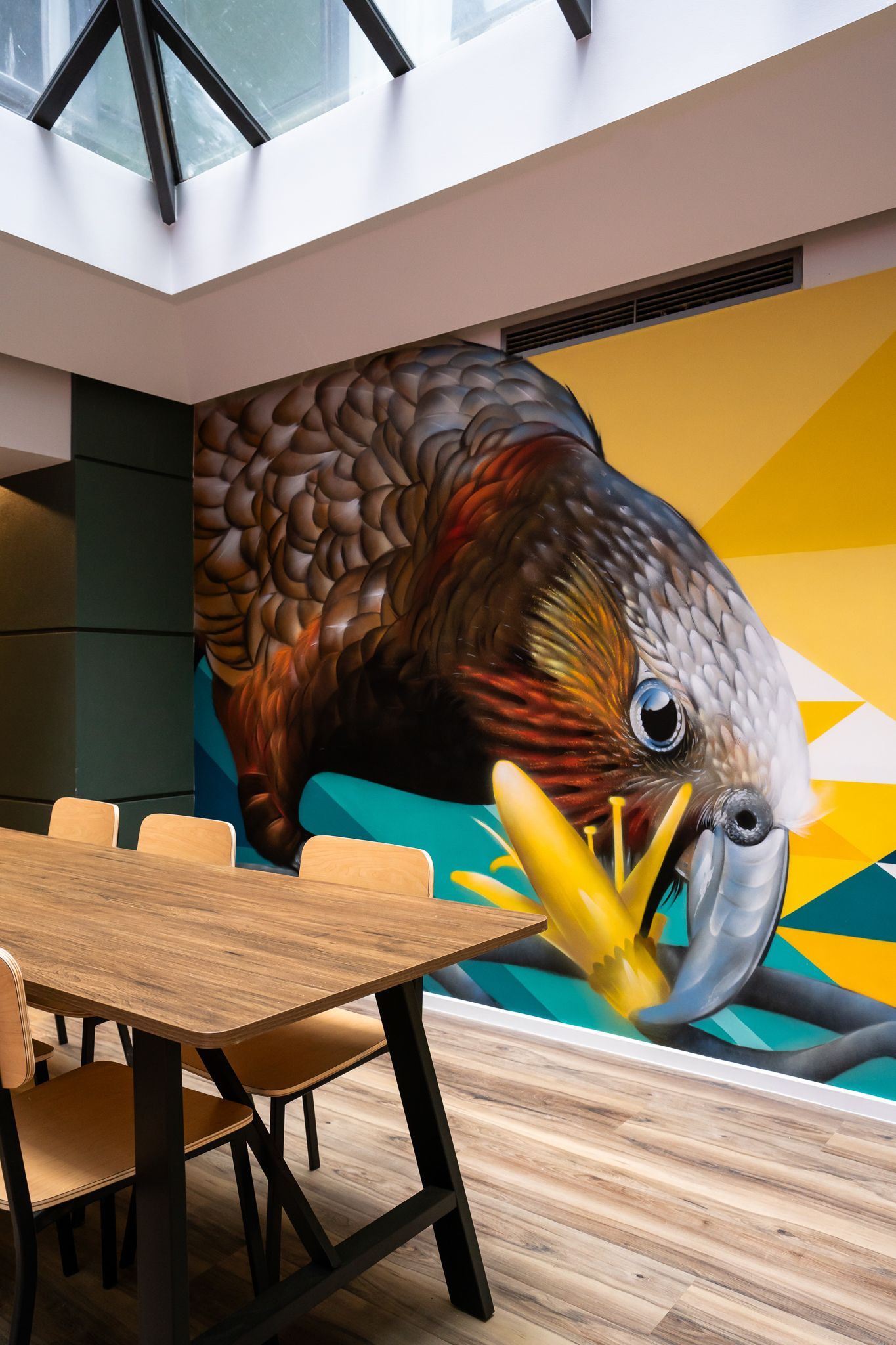Vibrant large-scale kea parrot mural in a bright dining area with wooden tables and chairs.