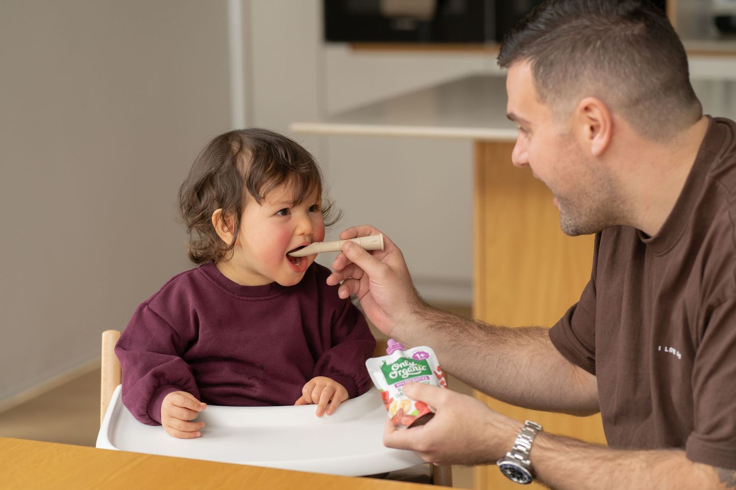 Father feeding toddler with a spoon at a high chair, sharing a happy mealtime moment together.