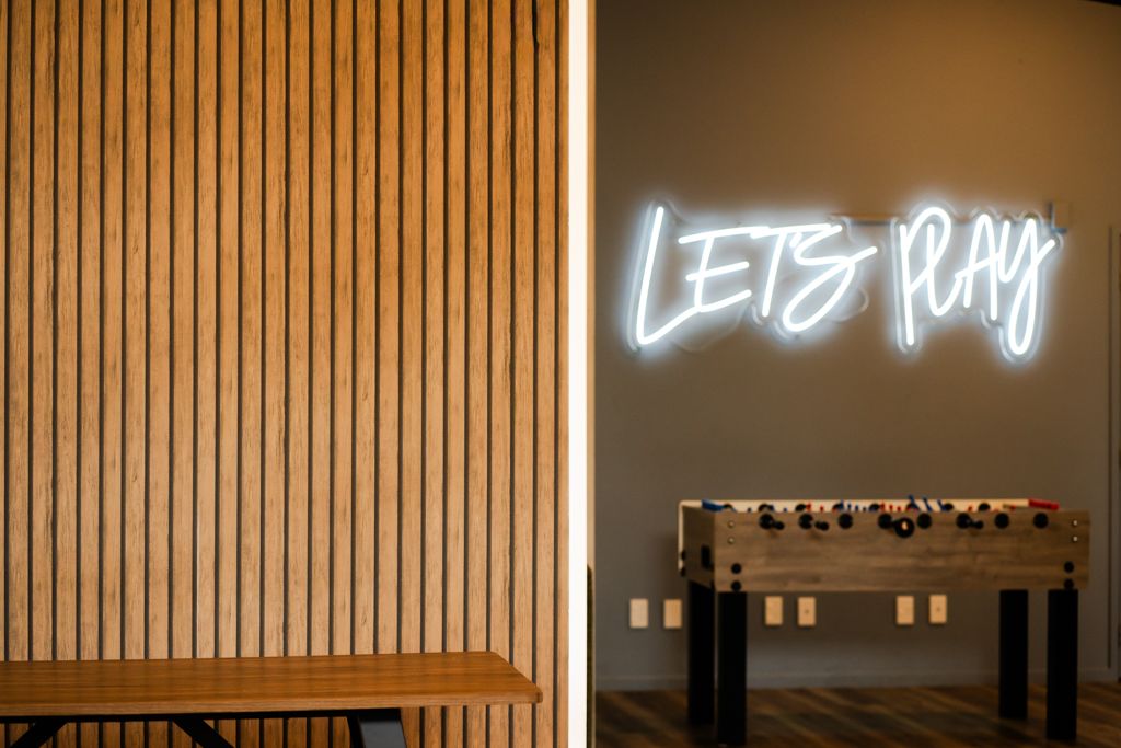 Wooden slat wall panel beside a glowing neon sign reading Lets Play above a foosball table.