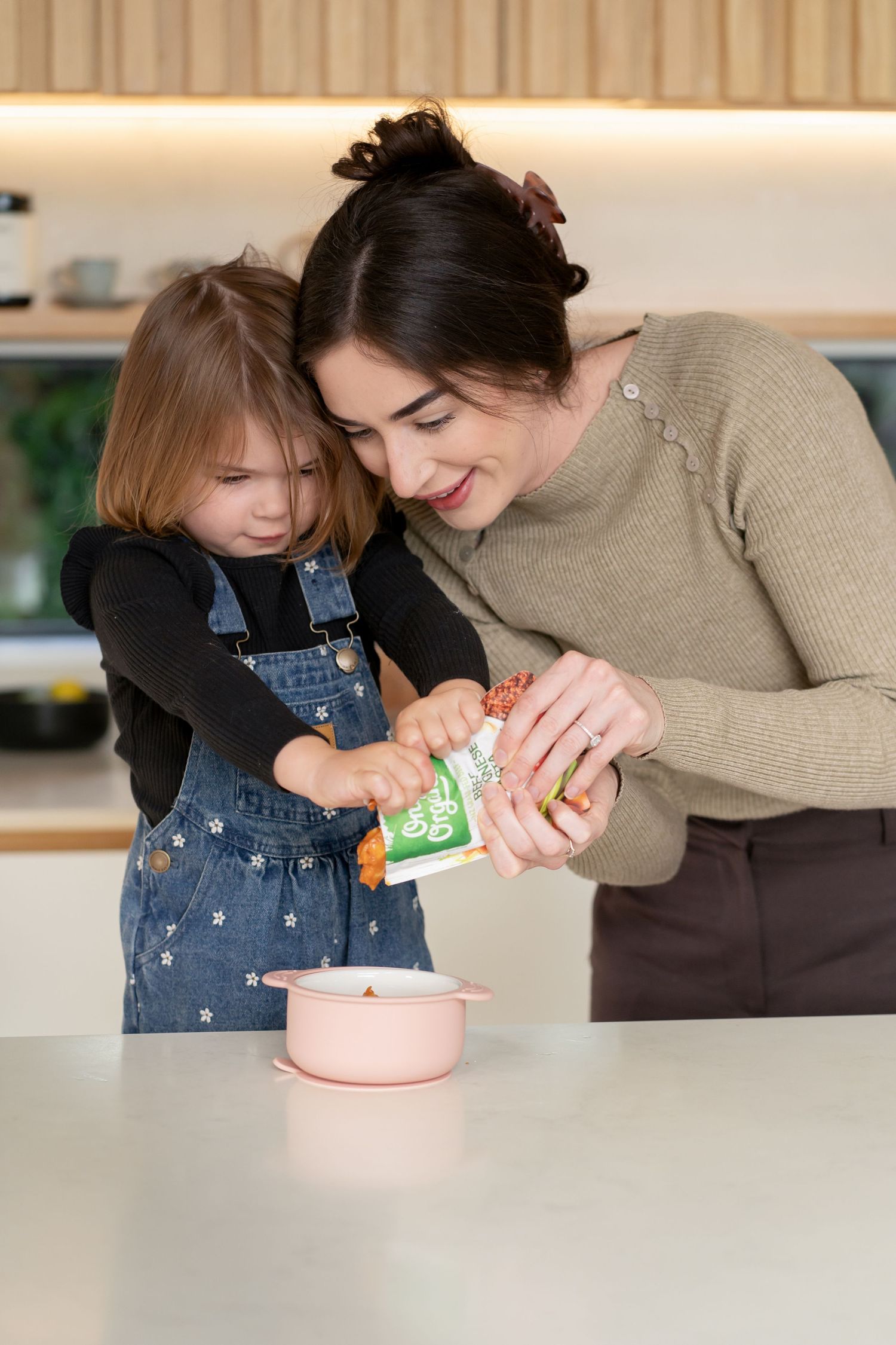 A smiling mother and young daughter cooking together in a modern kitchen, pouring ingredients into a pink pot.
