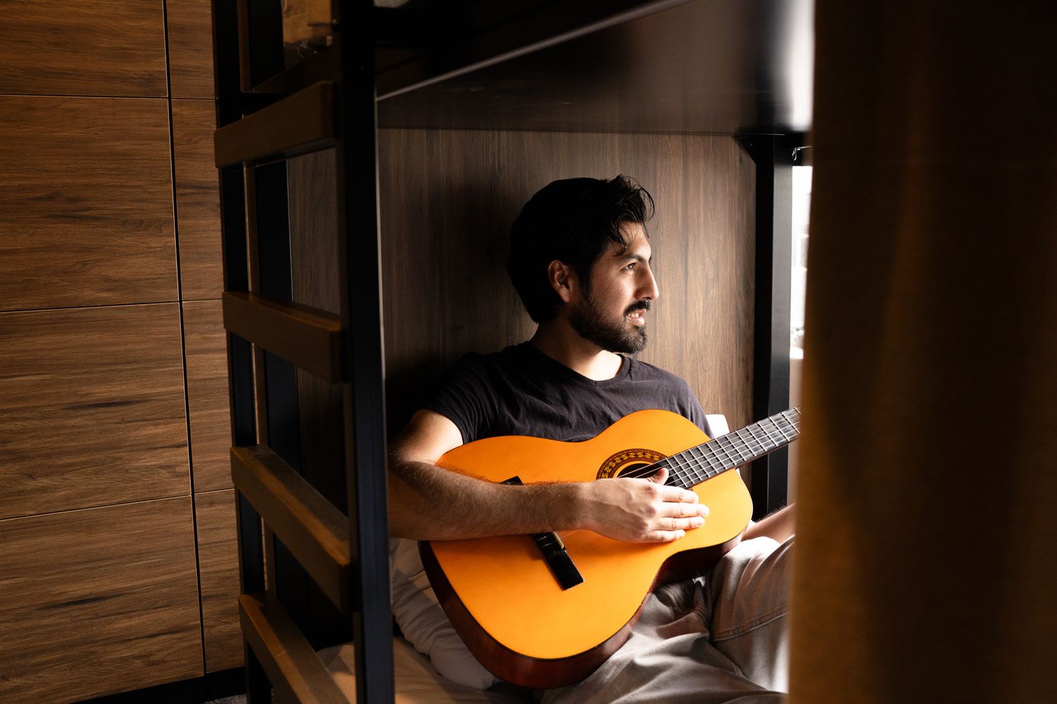 Bearded man playing acoustic guitar while sitting in a wooden hostel bunk bed pod.