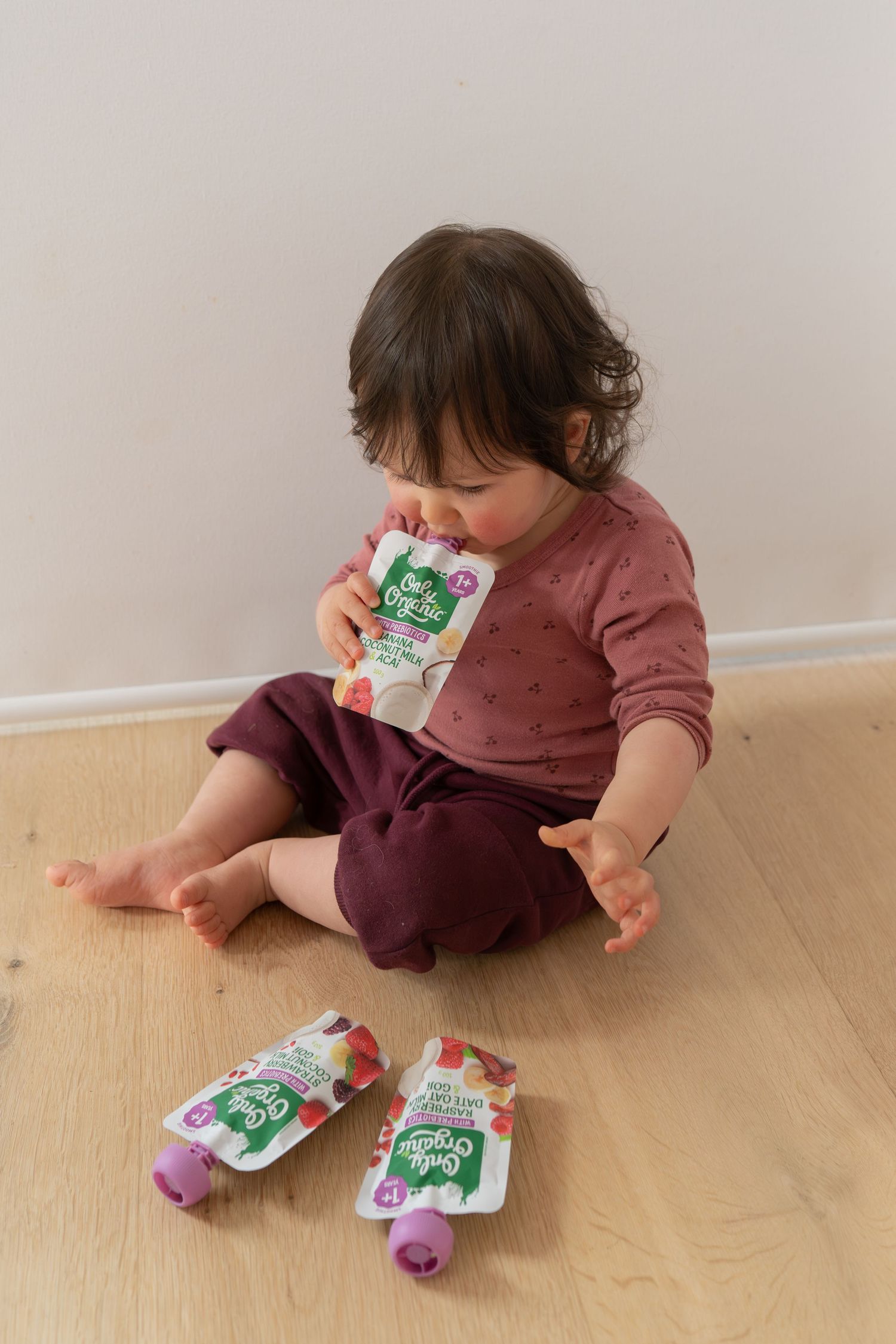 Toddler sitting on wooden floor eating from a Daily Organics fruit pouch, with two more pouches nearby.