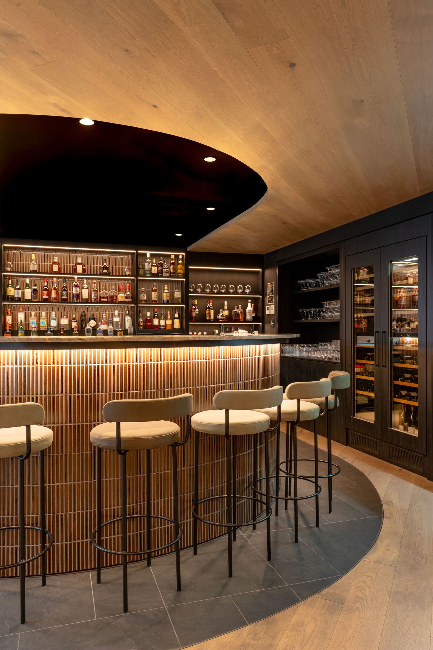 Sophisticated curved bar with warm timber details, cream upholstered stools, and illuminated bottle display.