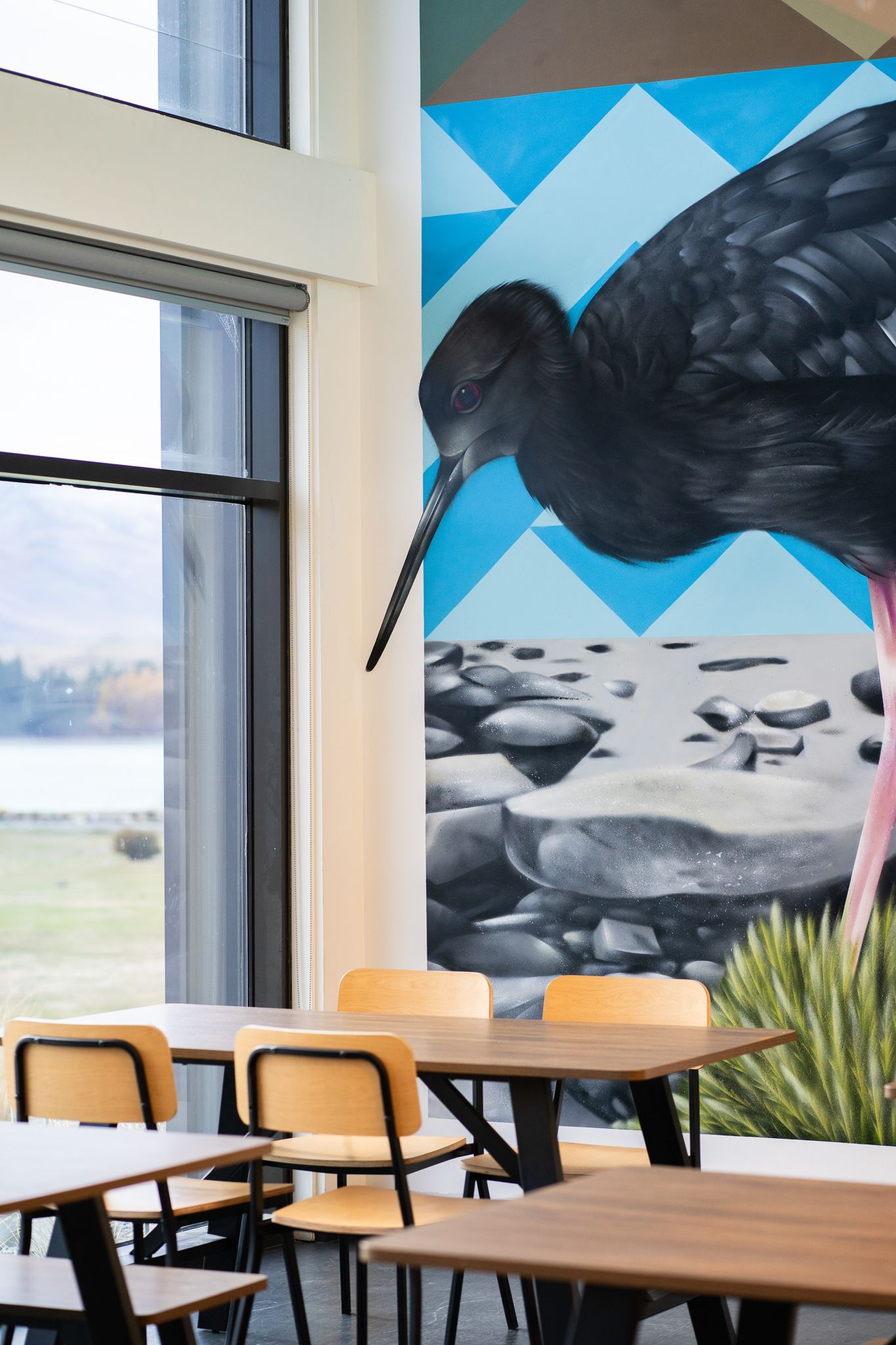 Dining area with wooden tables and chairs beside large windows featuring a bold black bird wall mural.