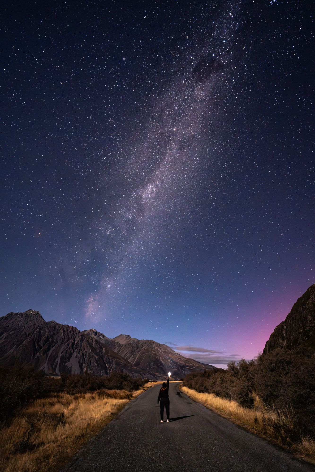 Person standing on road under stunning Milky Way galaxy night sky with mountains.